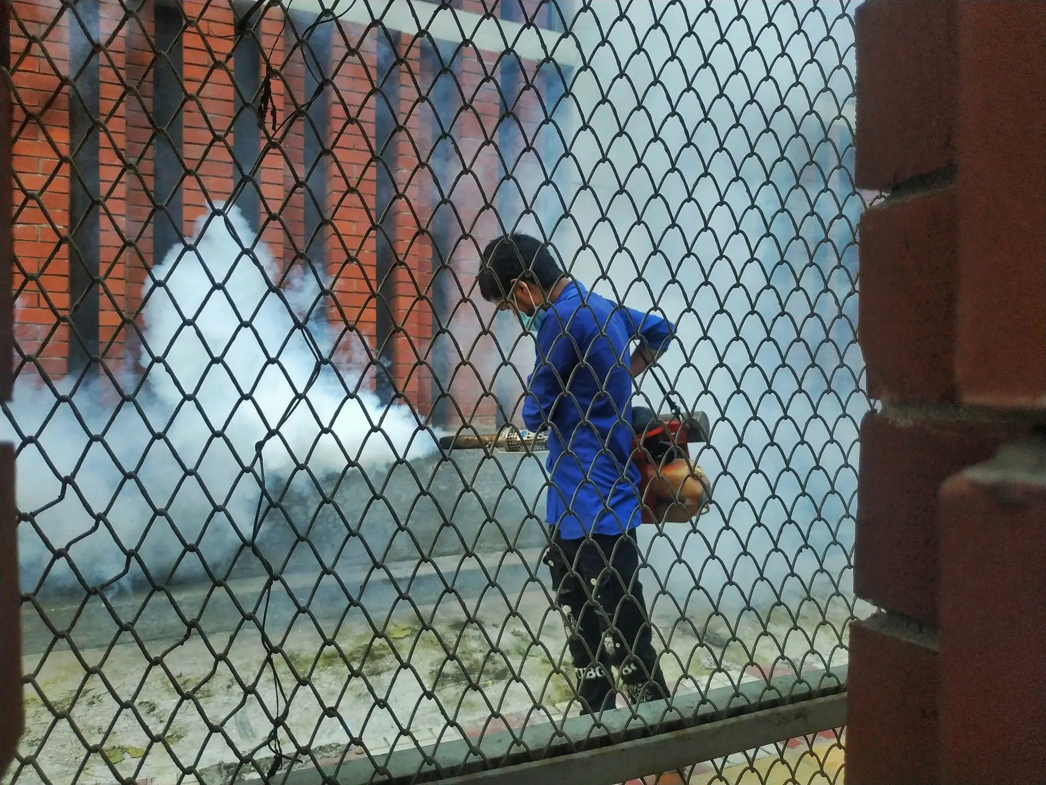 Person in blue uniform fogging for pests outside red brick building through wire fence
