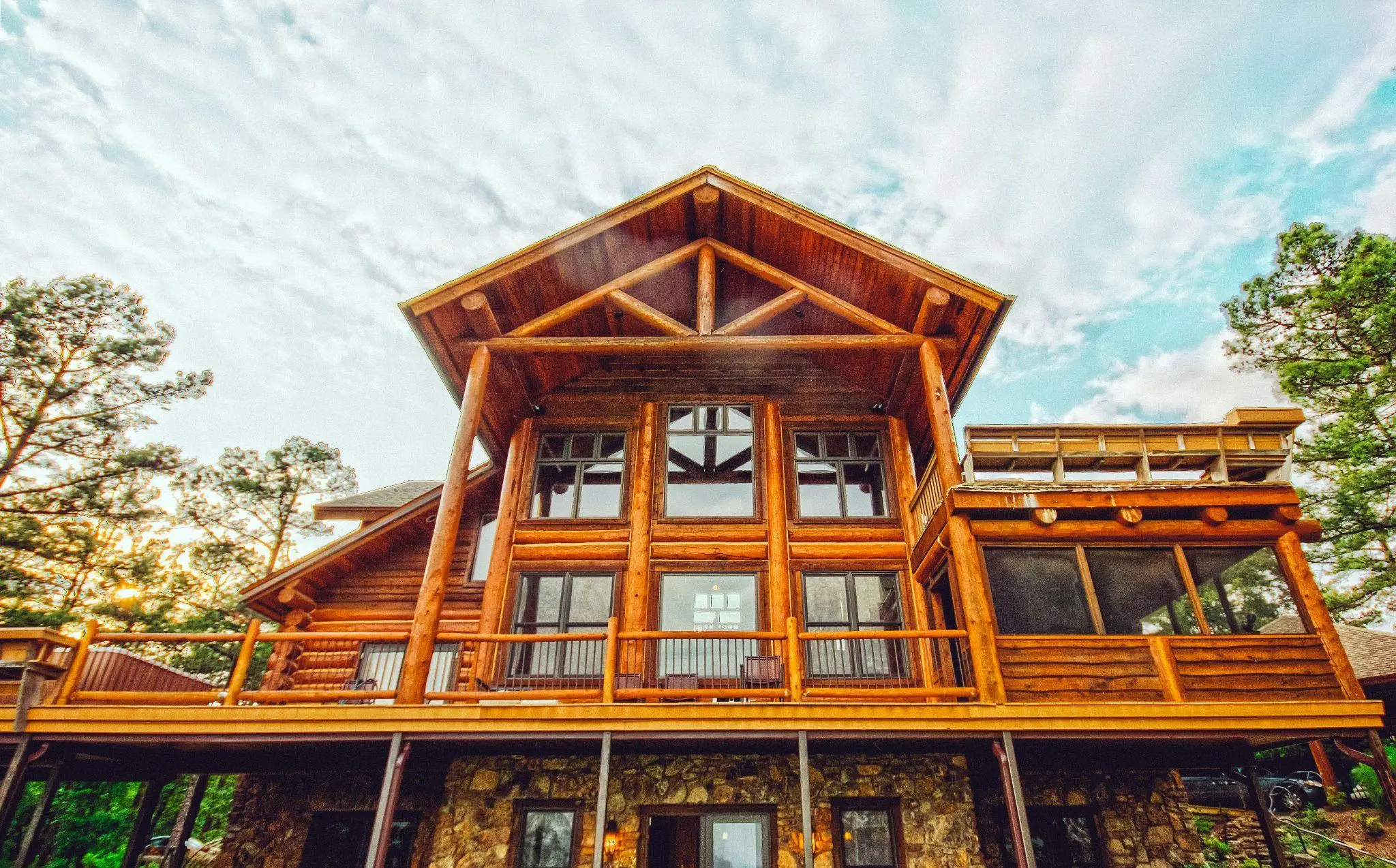 Large wooden log cabin with stone foundation surrounded by trees and cloudy sky