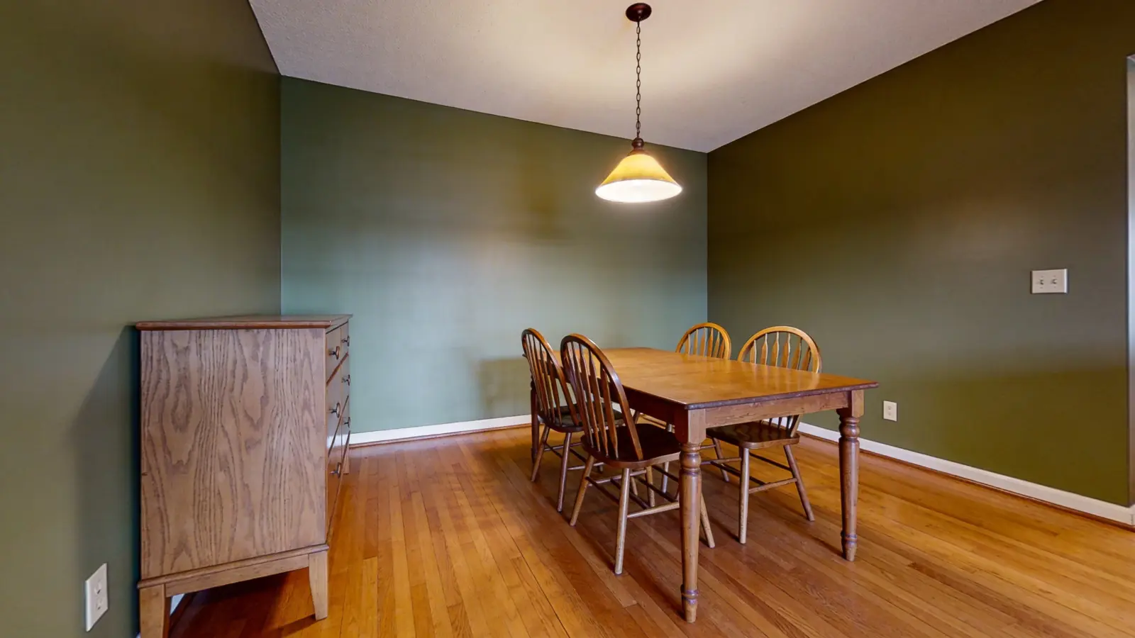 Wide view of dining room with dark green walls, wood table, chairs, and warm ceiling light