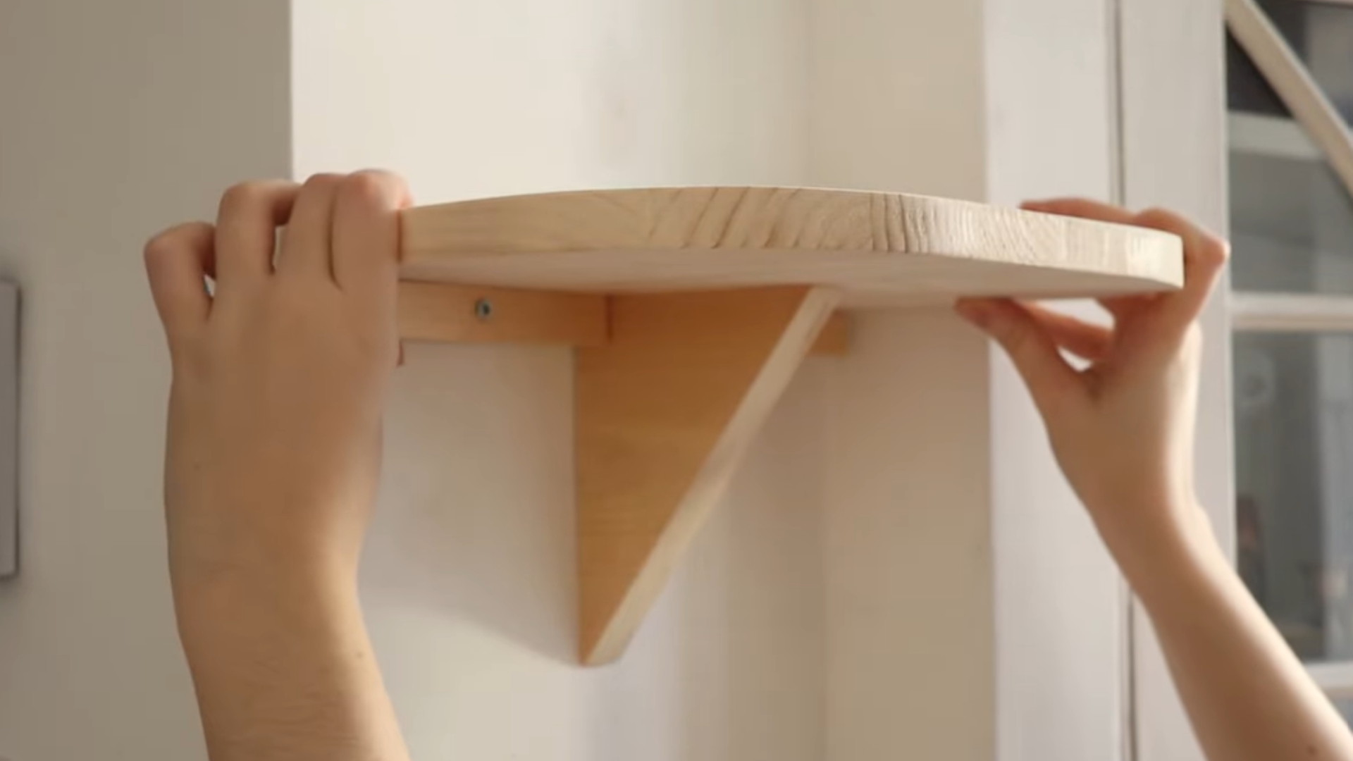 Hands installing a simple wooden corner shelf with triangular support on a white wall.