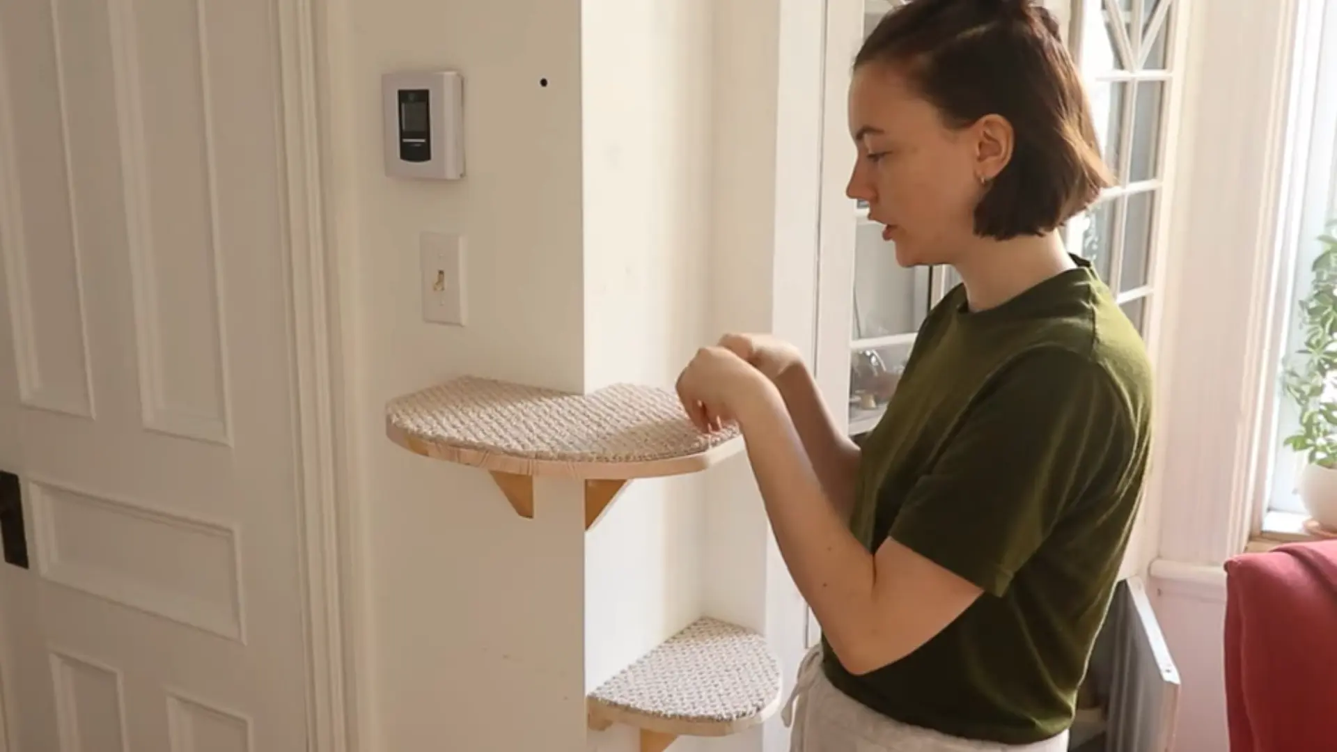 Person installing a carpeted corner cat shelf on a white wall near a doorway.
