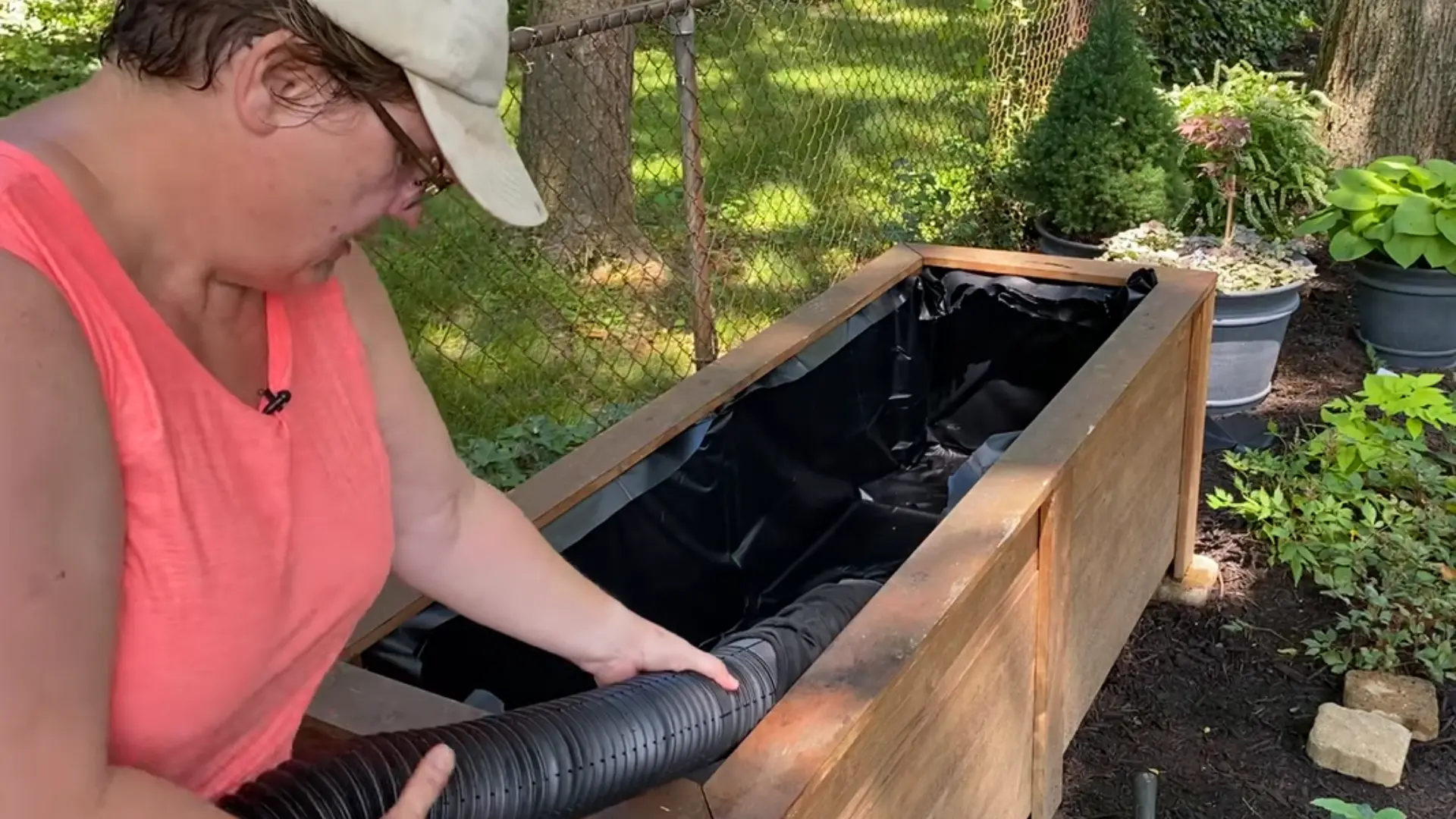 Person placing a flexible black drainage pipe inside a wooden planter lined with pond liner outdoors