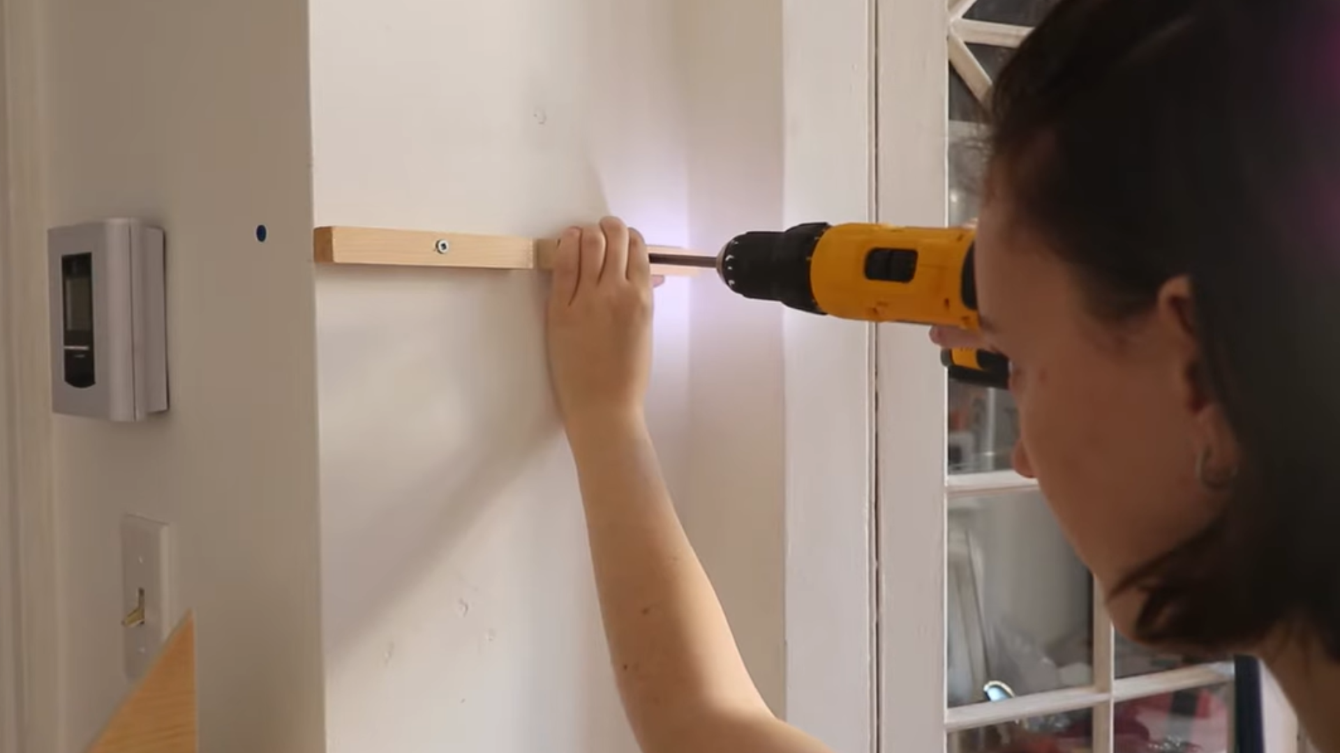 Person using a drill to attach a wooden bracket to a wall for mounting shelves.