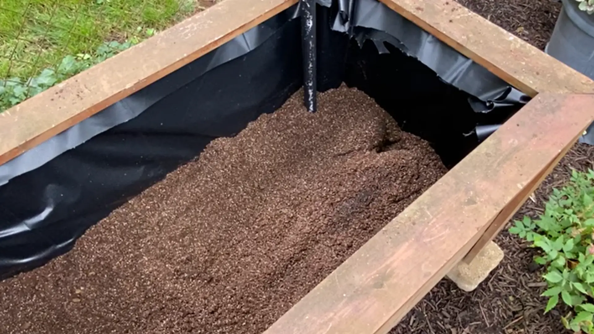 Wooden planter lined with black pond liner and partially filled with growing medium around a vertical pipe outdoors