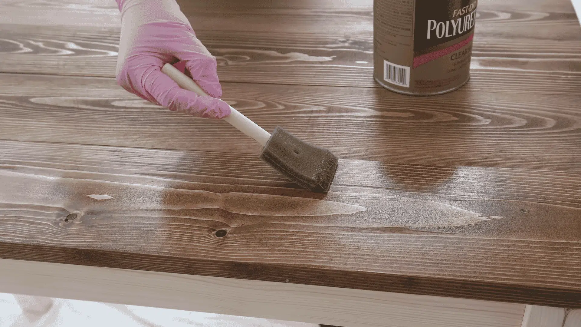 A gloved hand uses a foam brush to apply a clear polyurethane topcoat over a freshly stained wooden tabletop.