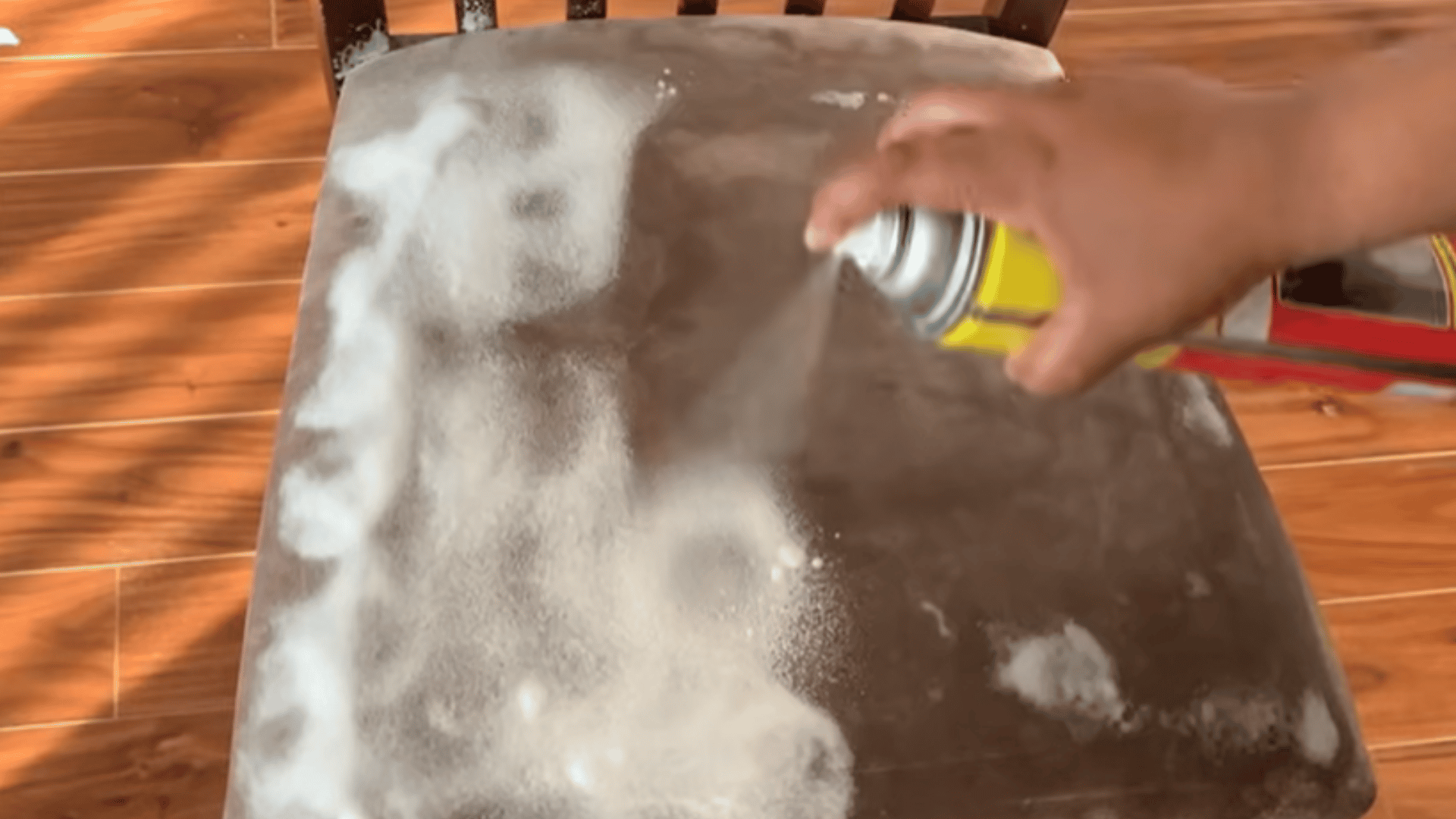 A hand spraying foam cleaner from a can onto a stained gray upholstered chair seat, with white foam spreading across the fabric. (1)