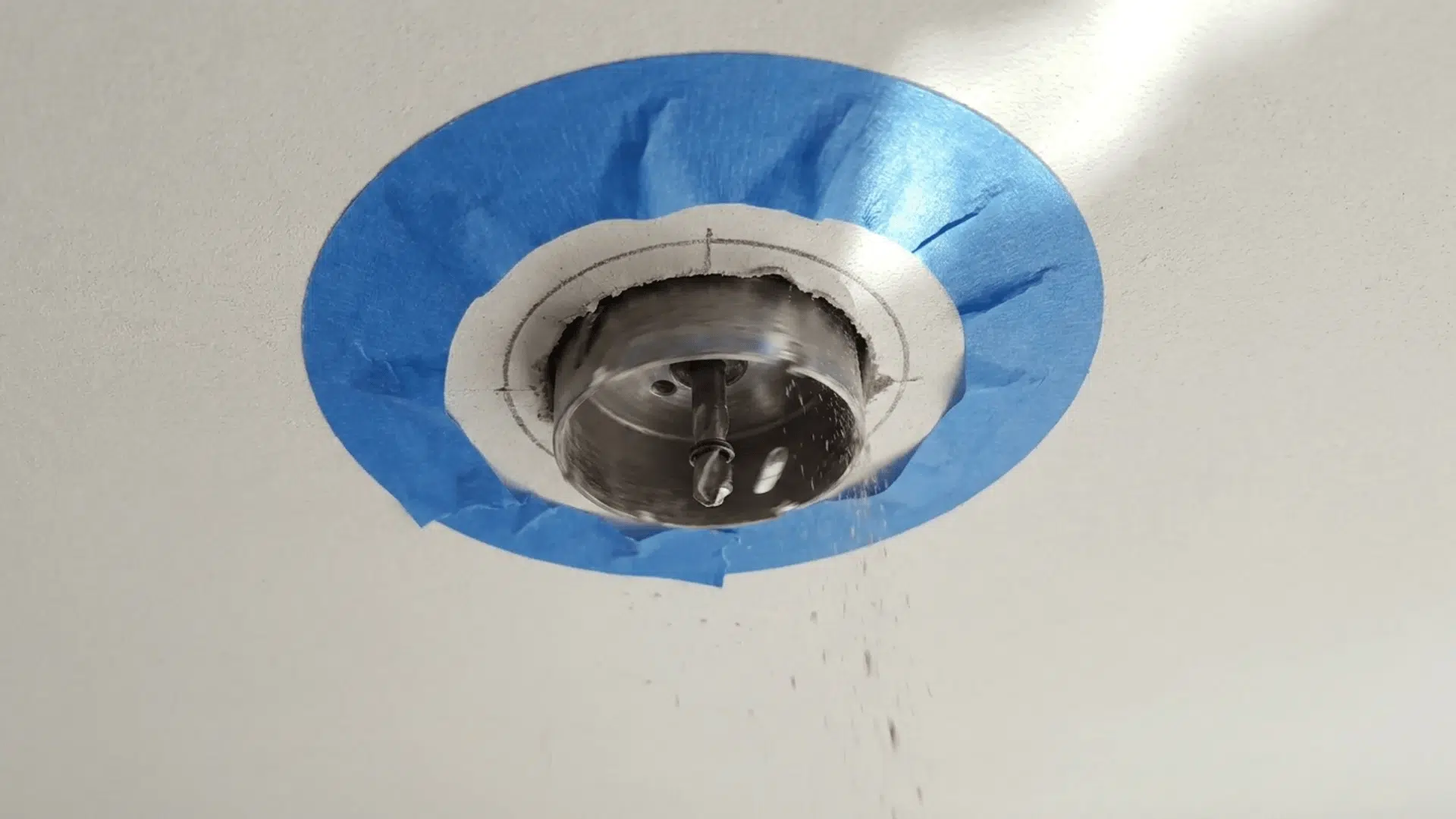 A hole saw drilling into the ceiling with painter's tape applied around the marked area to ensure clean, accurate cuts.