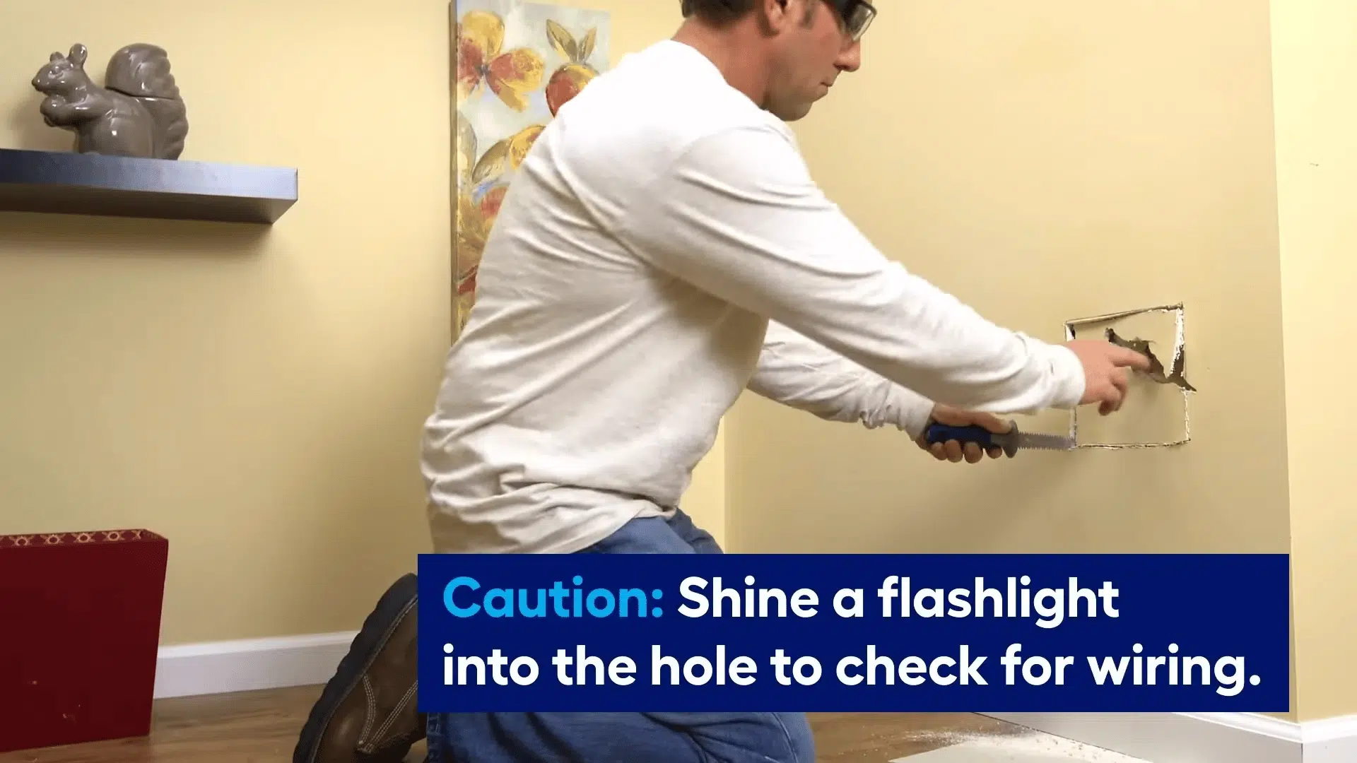 A man uses a drywall saw to cut a clean square around a damaged section of a wall while kneeling on the floor.