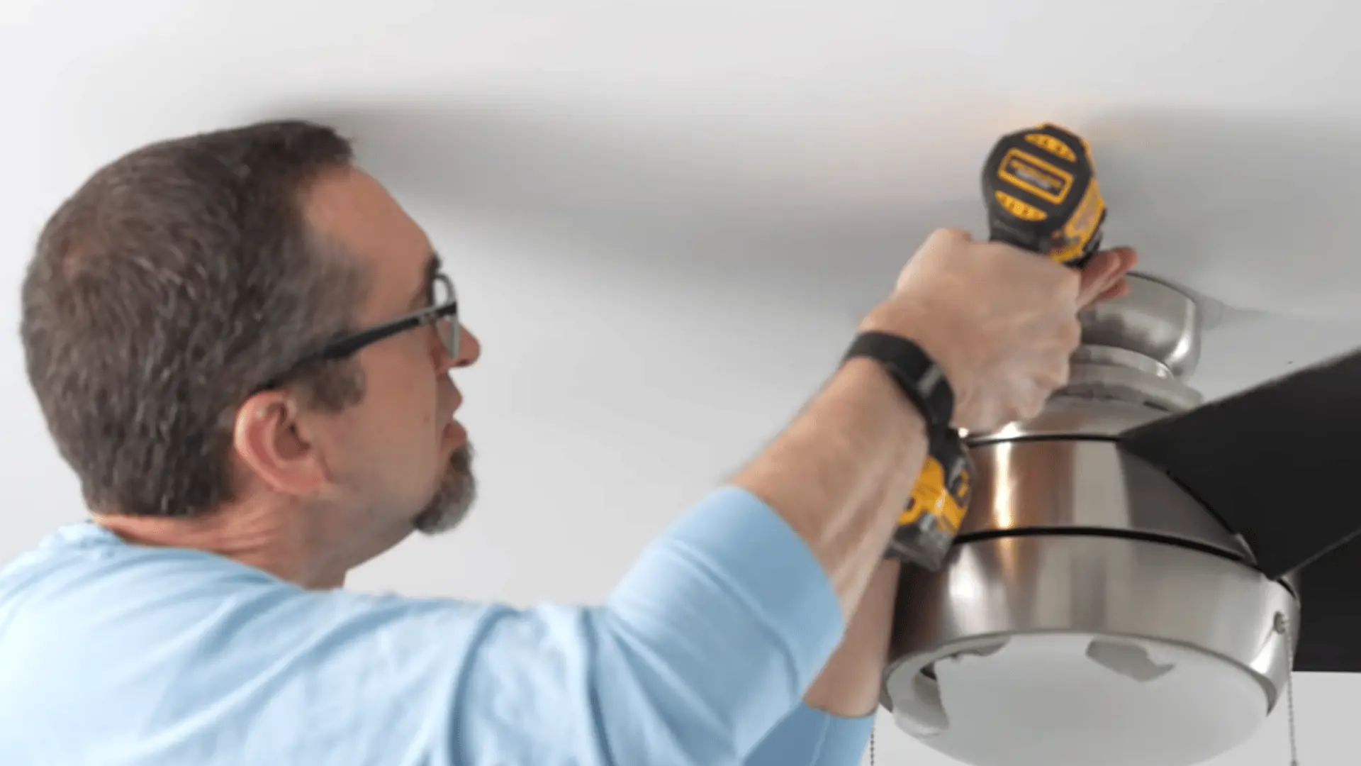 A person attaching the ceiling fan motor housing to the ceiling using a drill during installation.