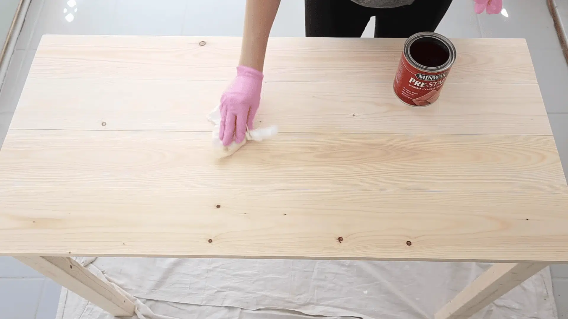 A person in pink gloves wipes liquid pre-stain wood conditioner onto a light wooden tabletop using a white rag.