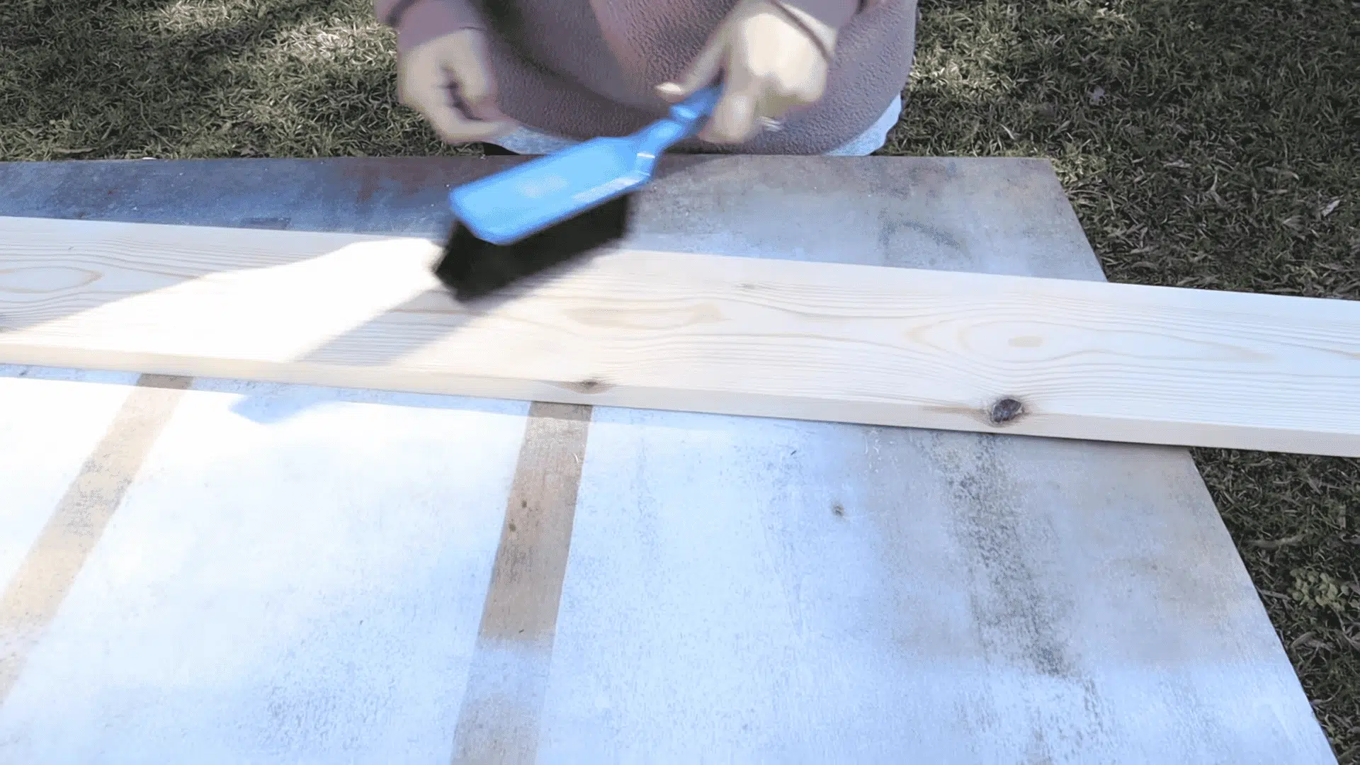 A person uses a blue brush to sweep sawdust off a long, light-colored wooden board outdoors.