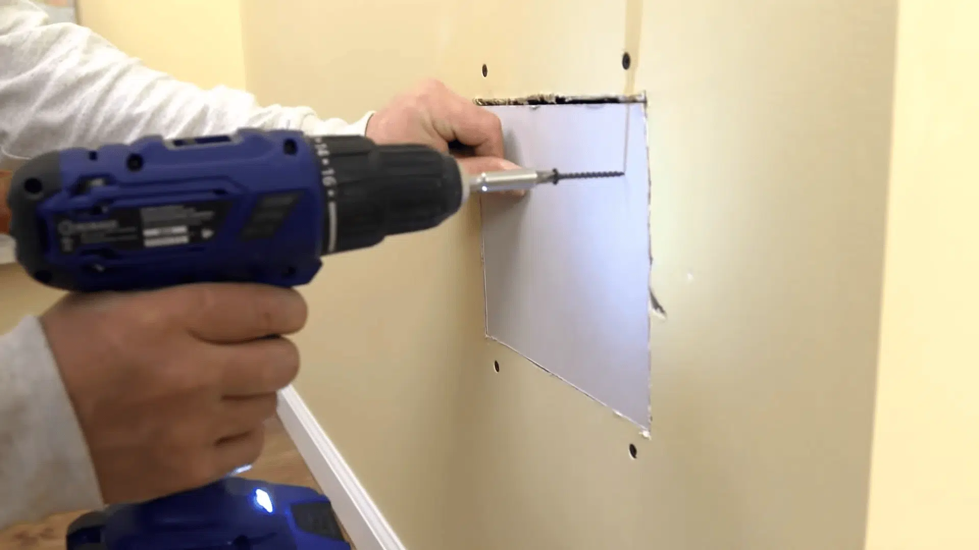 A person uses a blue power drill to drive a screw into a square drywall patch, securing it to the wall.