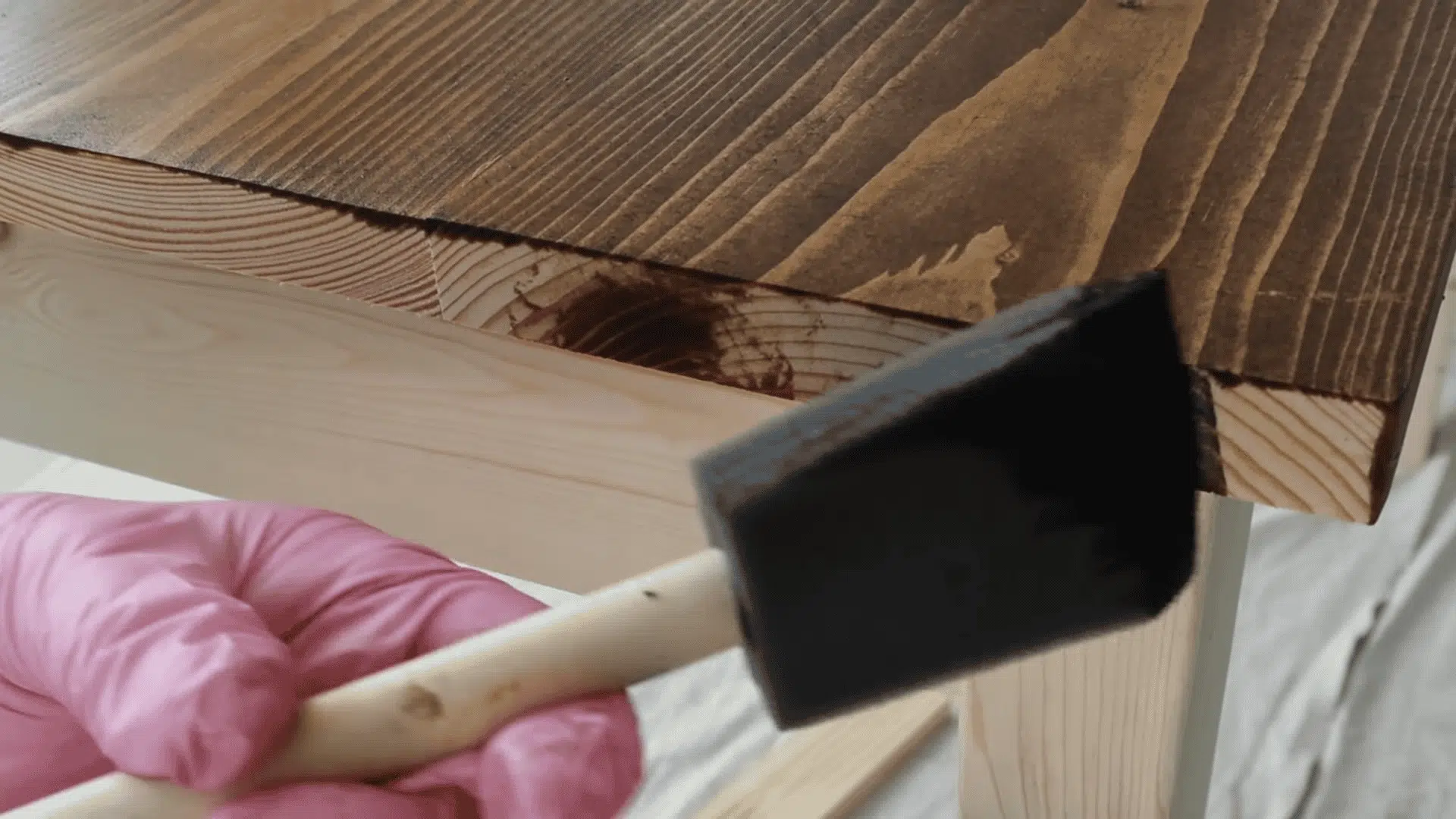 A person uses a foam brush to apply dark wood stain to the side edge of a light-colored wooden tabletop.