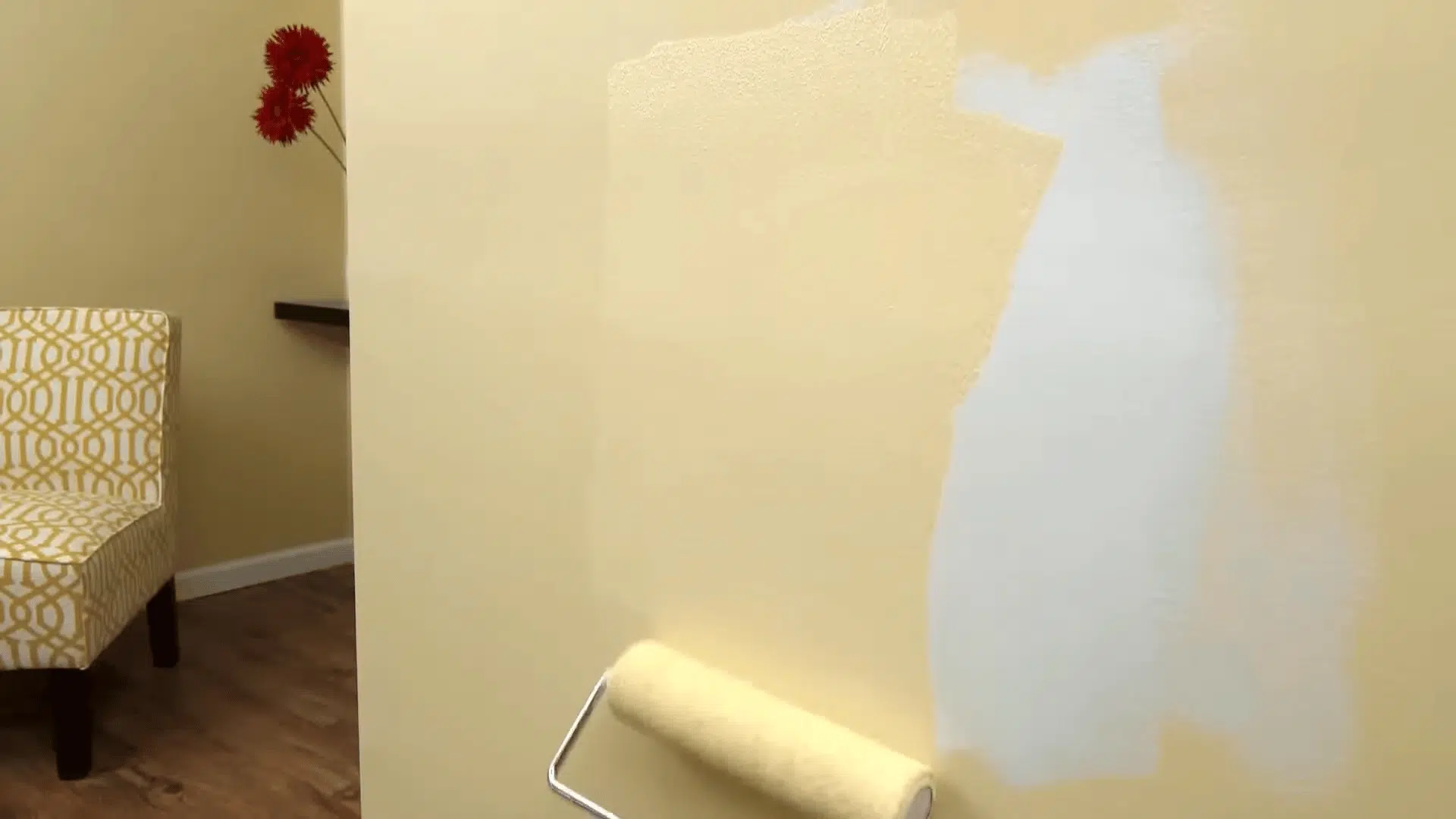 A person uses a paint roller to apply a tan-colored coat over a white primed section of a wall.
