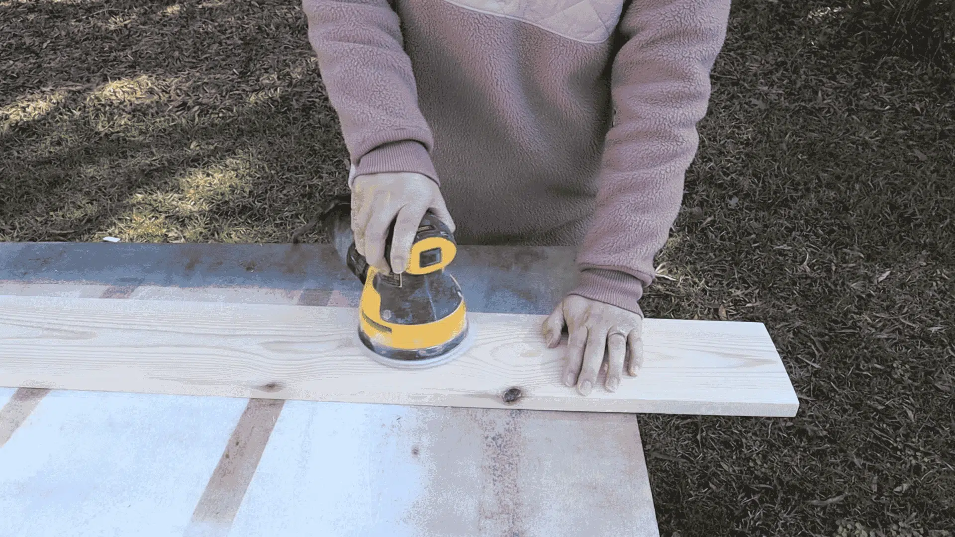A person uses a yellow orbital sander to smooth the surface of a long pine board placed on a workbench outdoors.