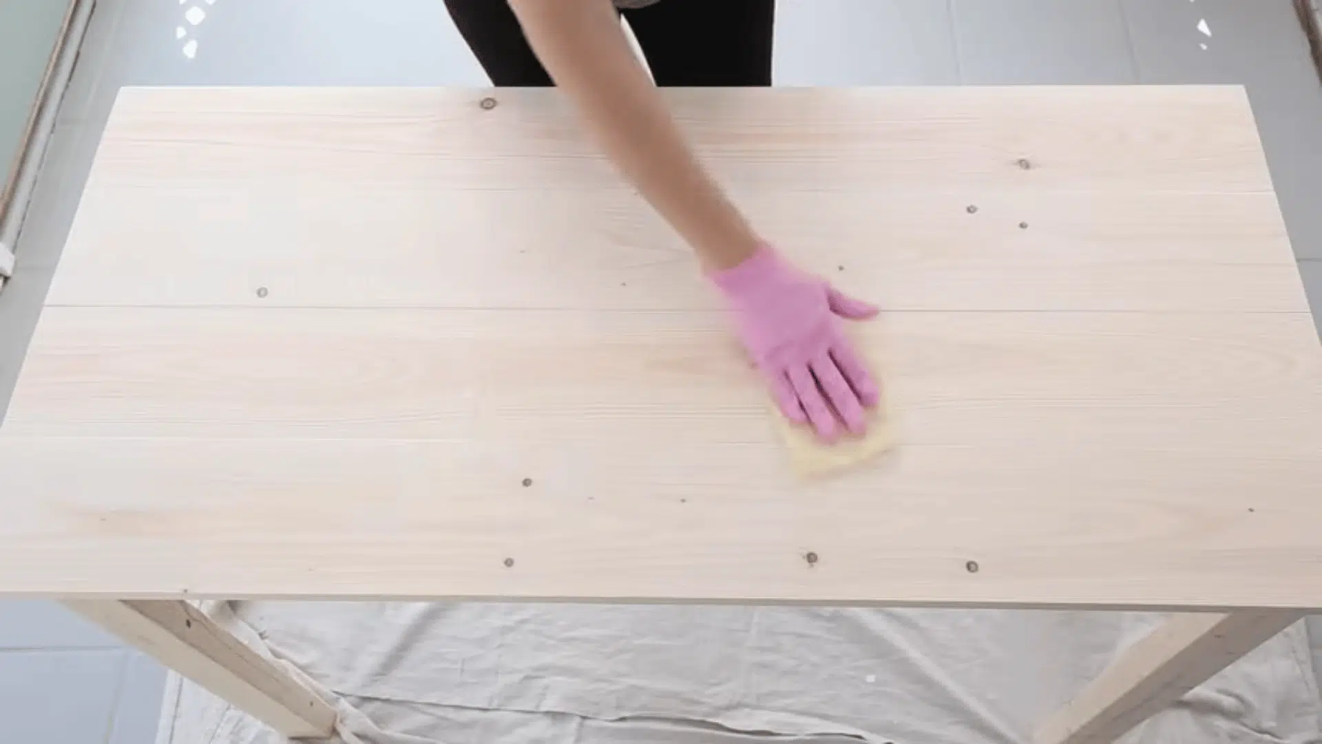 A person uses a yellow tack cloth to wipe down a light wood tabletop, removing fine dust particles before staining.