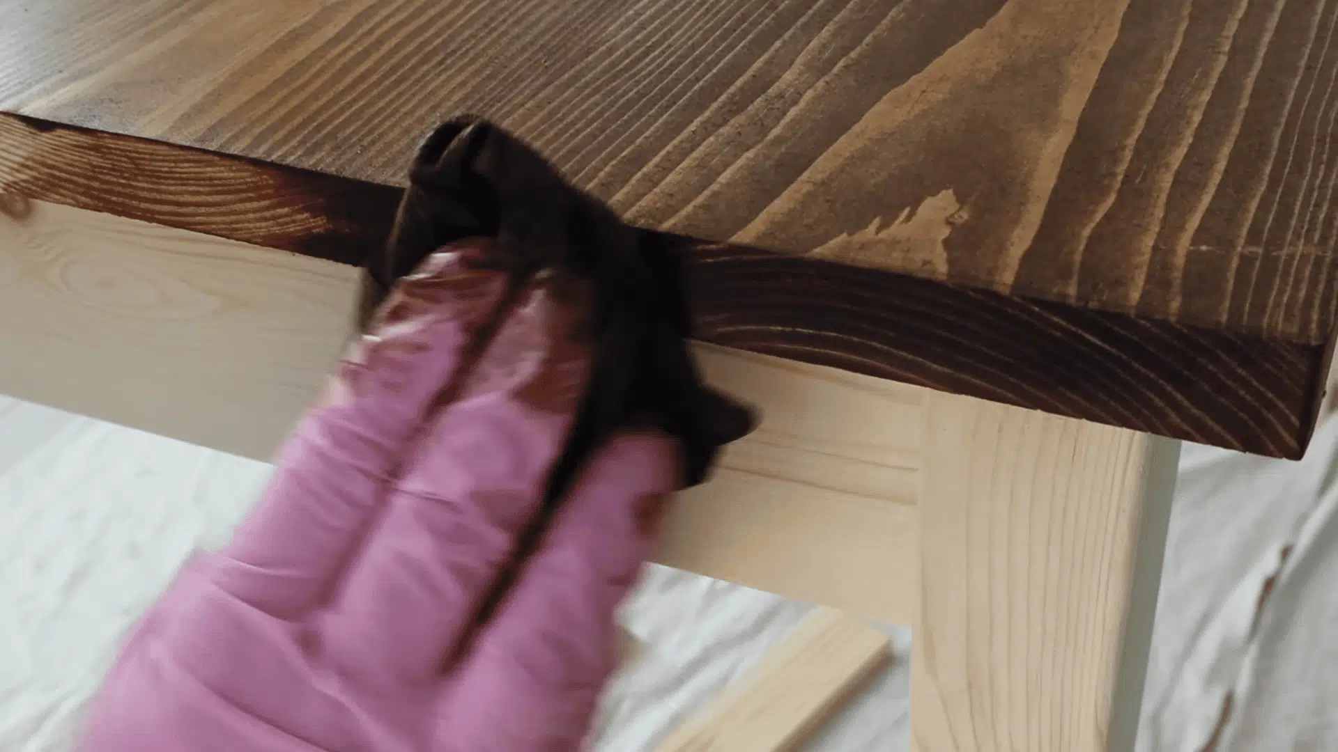 A person wearing pink gloves uses a dark lint-free rag to wipe excess stain off the edge of a wooden table.