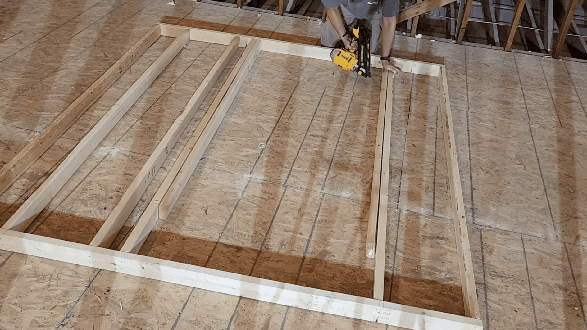 carpenter securing wall studs with a framing nailer while assembling a wooden wall frame layout on the subfloor during construction