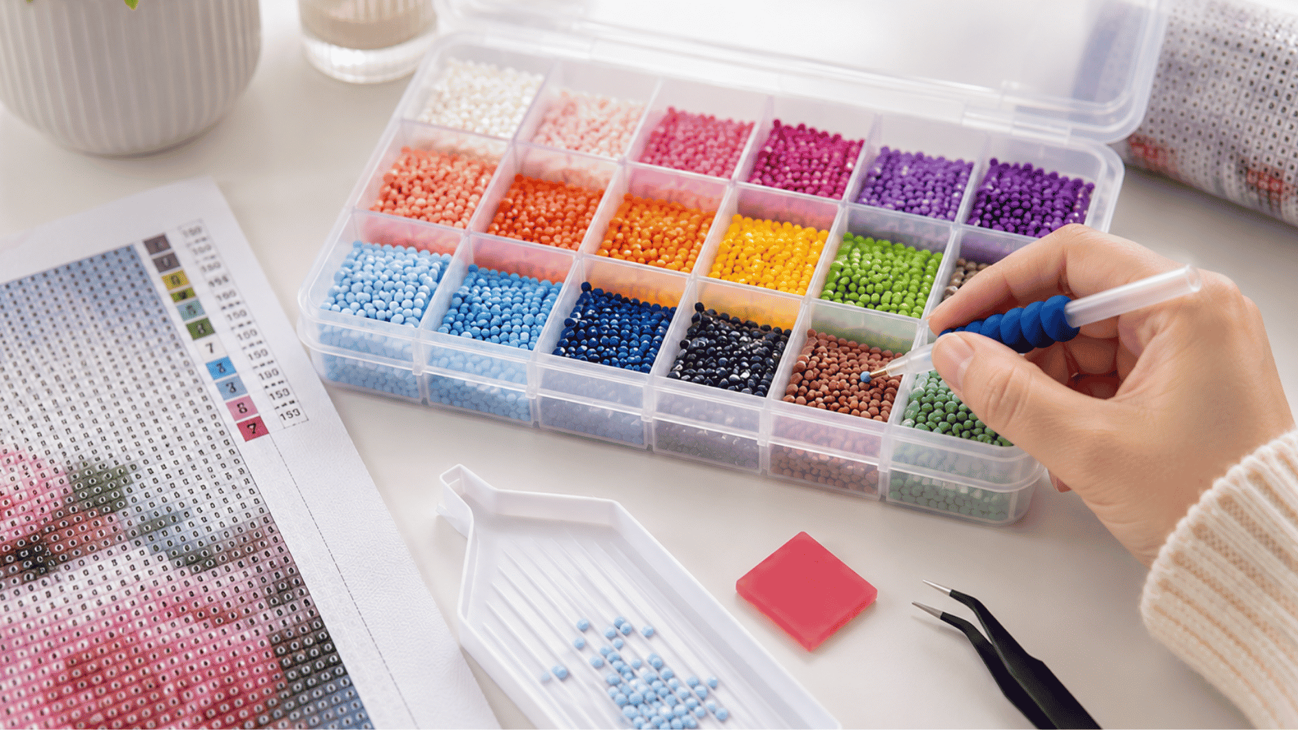 close-up of a hand organizing colorful resin diamonds in a storage container with diamond painting tools, tray, wax pad, and canvas on a clean crafting table under soft lighting