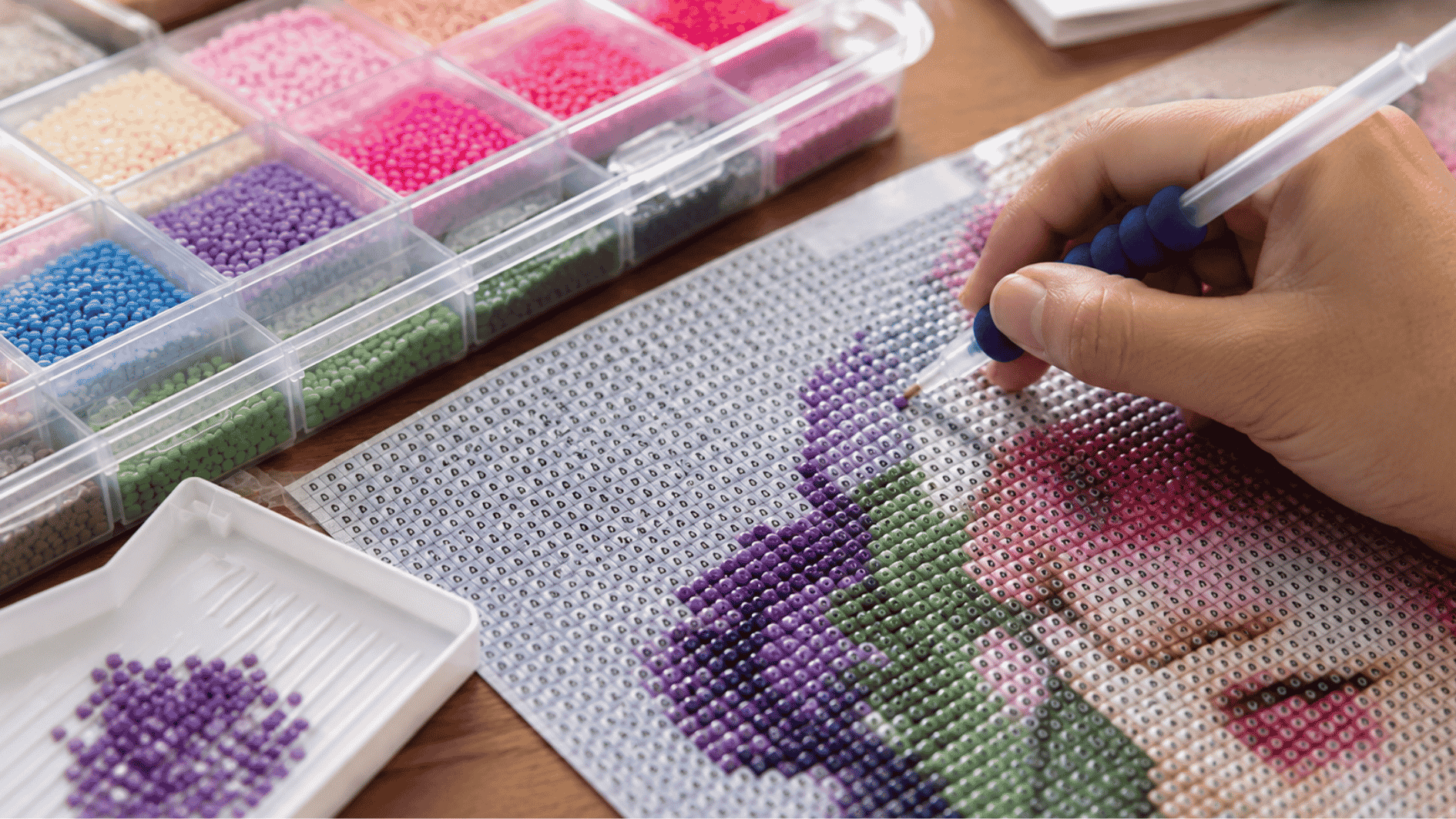 close-up of a hand placing colorful resin diamonds onto matching symbols on a diamond painting canvas with organized trays, applicator pen, and crafting tools on a wooden table under soft indoor l