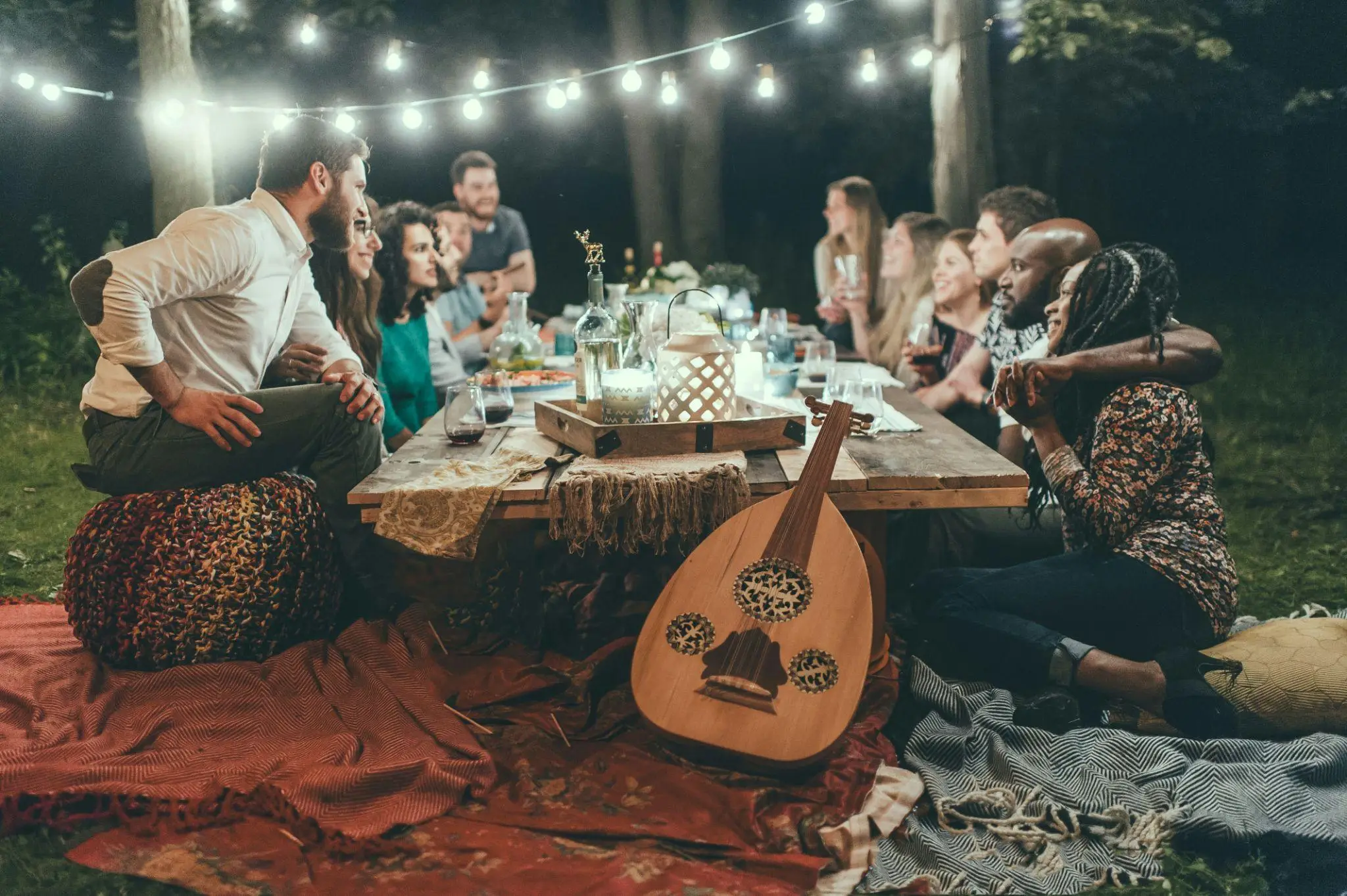 Group of people enjoying an outdoor dinner party with string lights and musical instrument