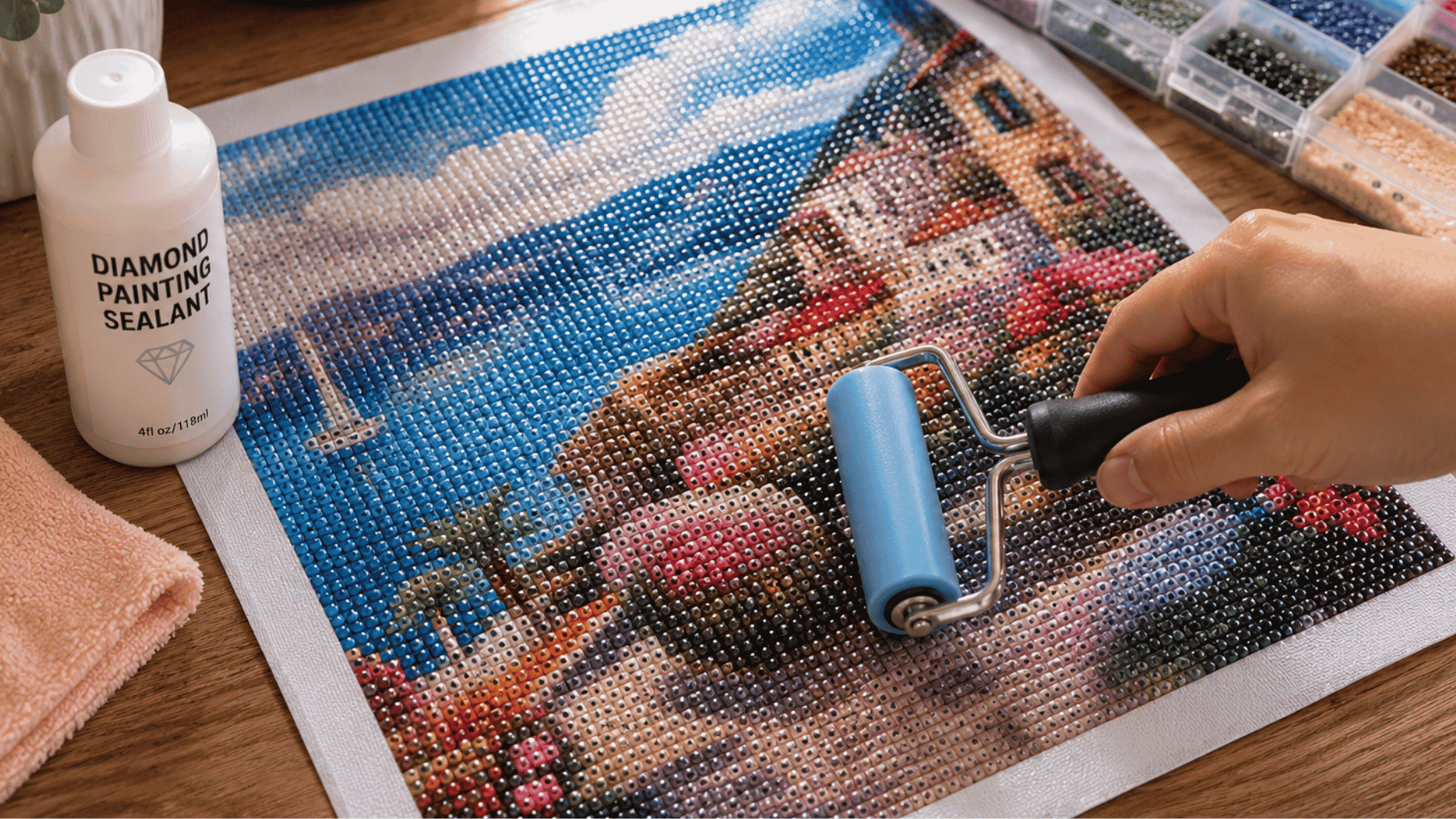 finished diamond painting artwork being pressed evenly with a roller on a wooden crafting table beside sealant and colorful resin diamonds