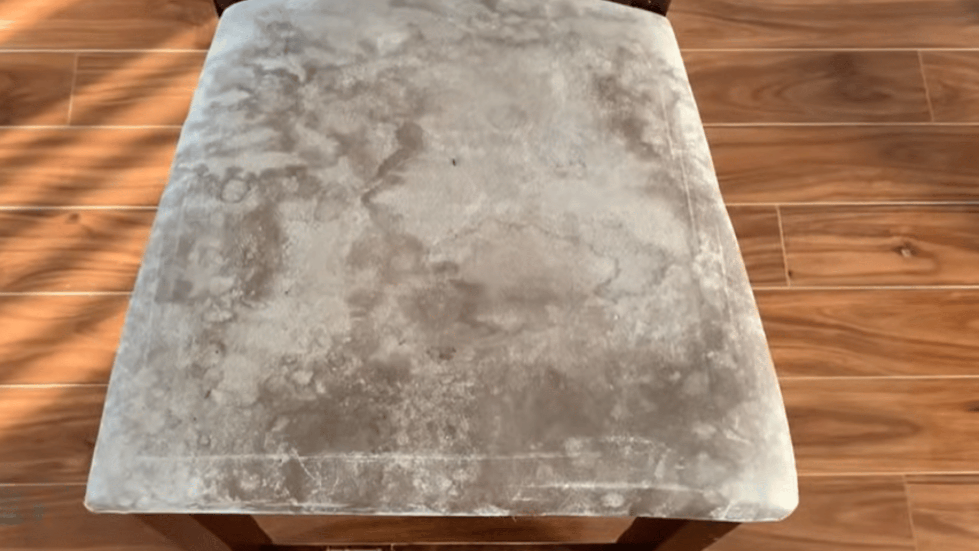 Gray upholstered chair seat with uneven light and dark stains, set on a wooden floor in sunlight.