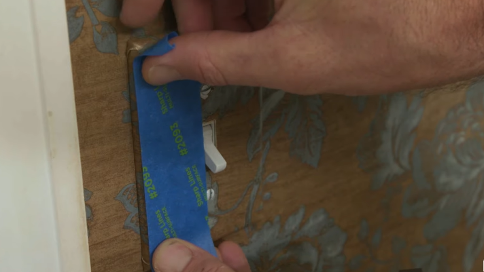Hand applying blue painter’s tape around a light switch plate on a patterned wall