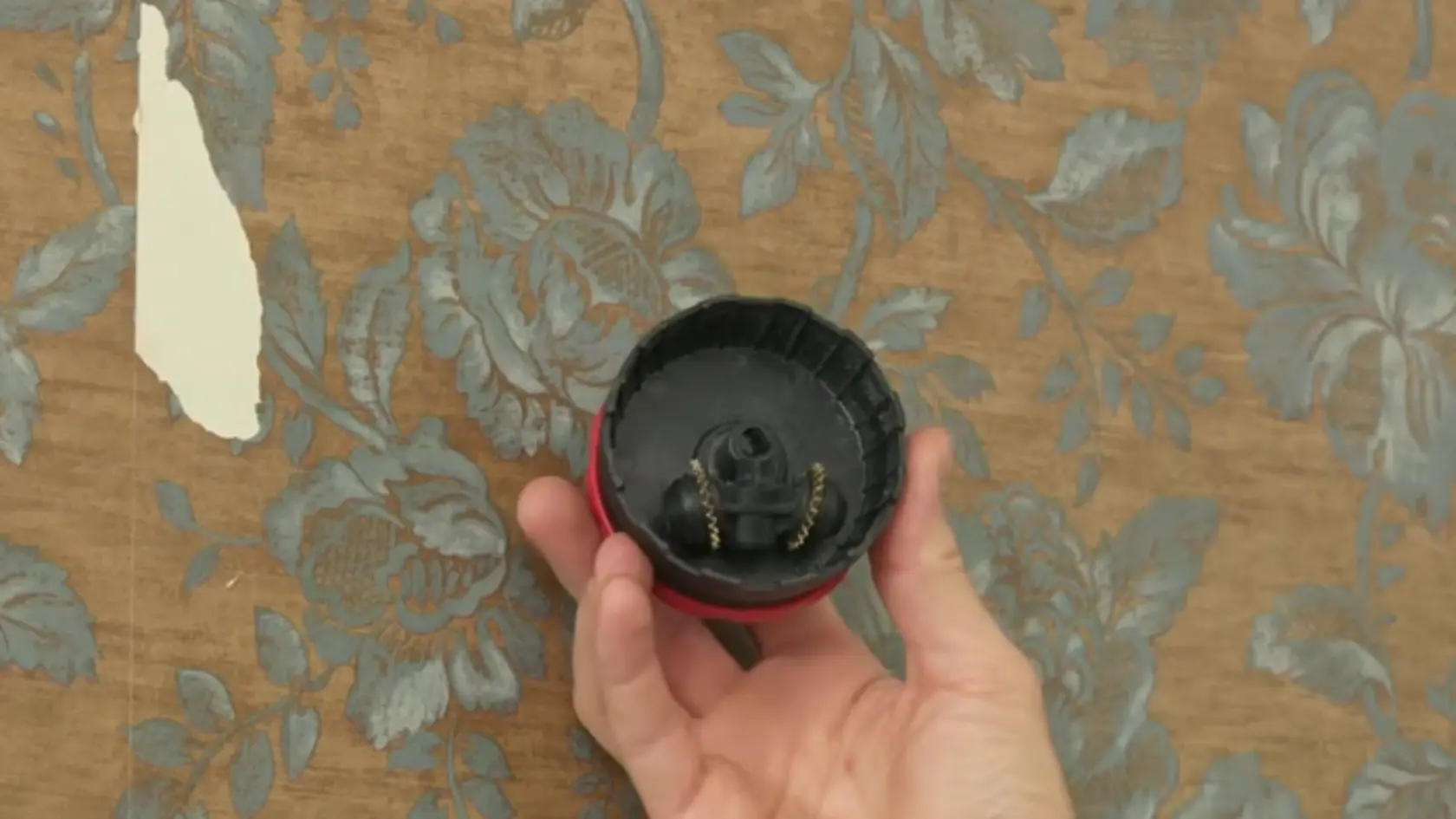 Hand holding a hole saw drill attachment in front of partially removed floral wallpaper on a wall