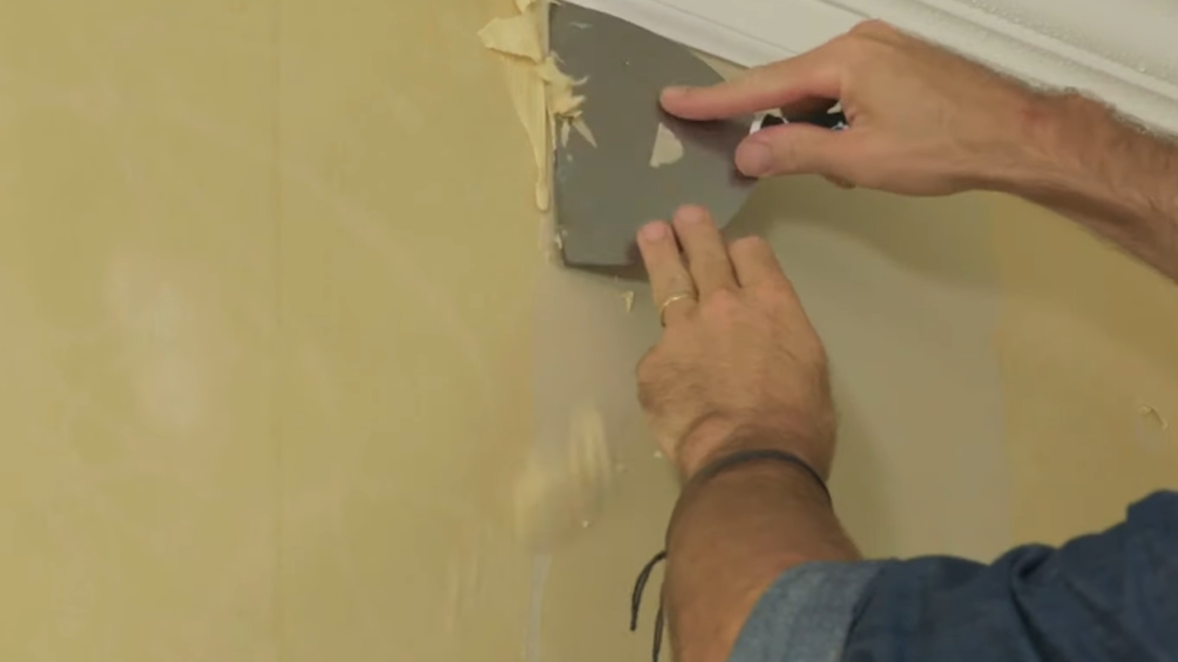 Hand using a scraper to remove softened wallpaper near ceiling trim