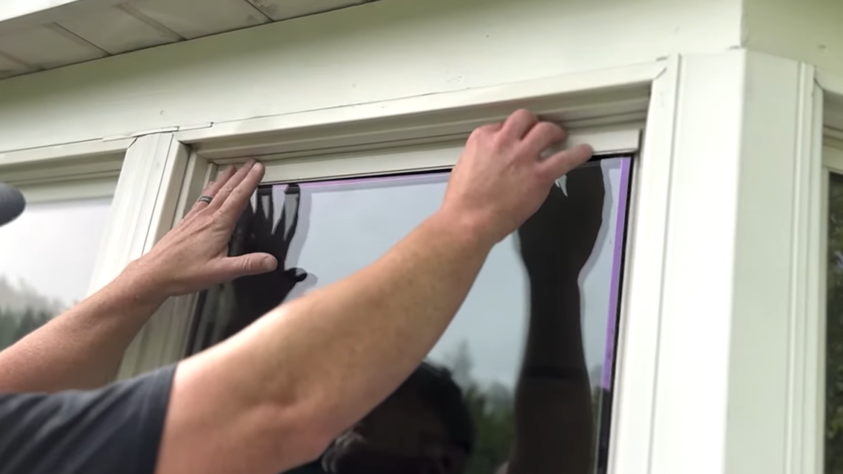 Hands positioning a replacement window pane into a white exterior window frame outdoors