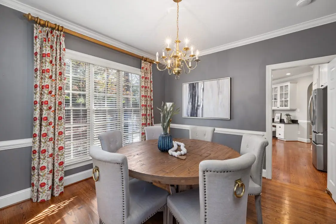 Dining room with round wooden table, gray chairs, floral curtains, and gold chandelier in bright setting