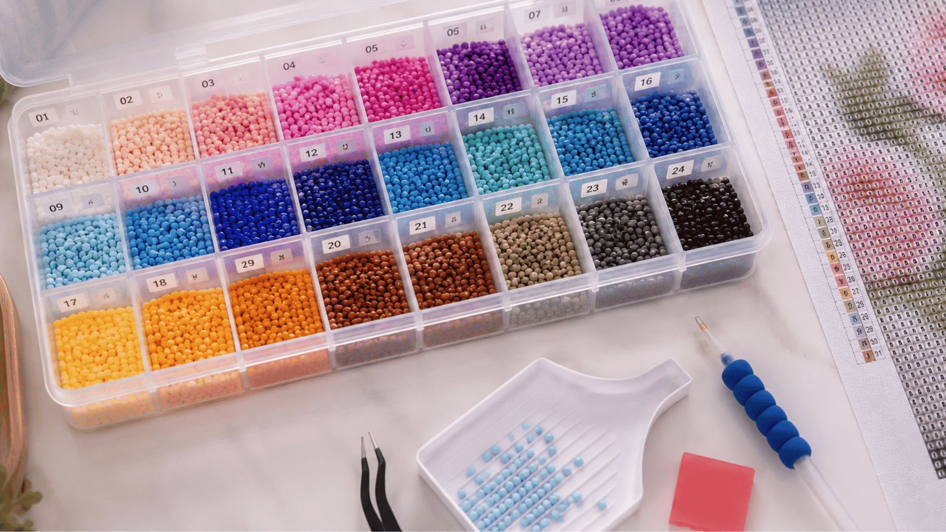 organized diamond painting setup with colorful resin diamonds sorted by color in labeled storage compartments beside a canvas, tray, applicator pen, tweezers, and wax pad on a crafting table