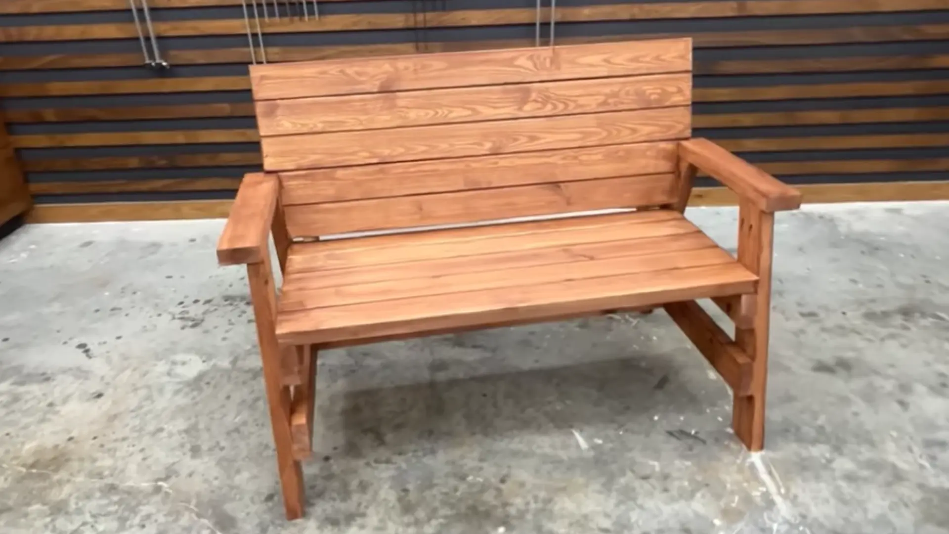 Person-built wooden bench with slatted seat and back, finished in a warm stain, standing on a concrete floor in a workshop