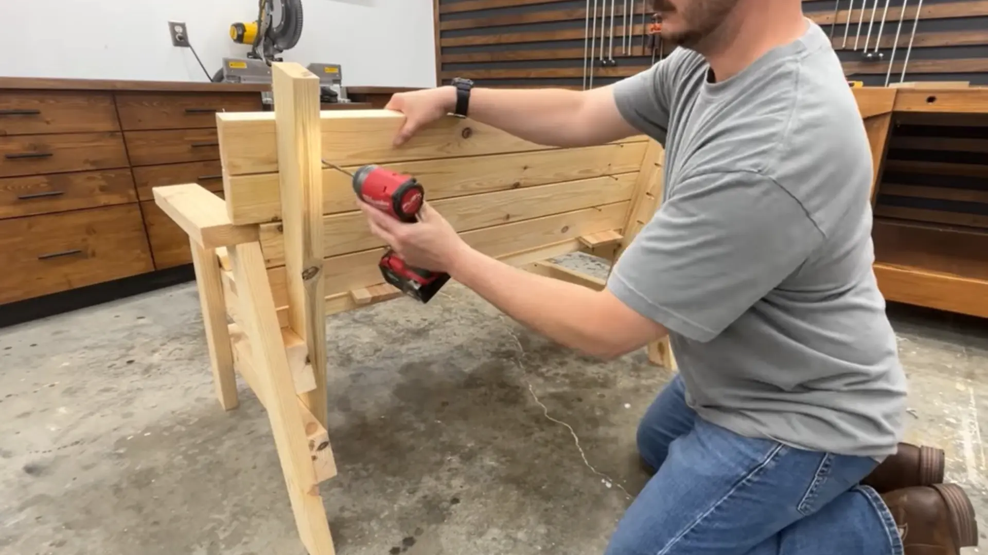 Person kneeling in a workshop, drilling screws into wooden frame for furniture assembly.