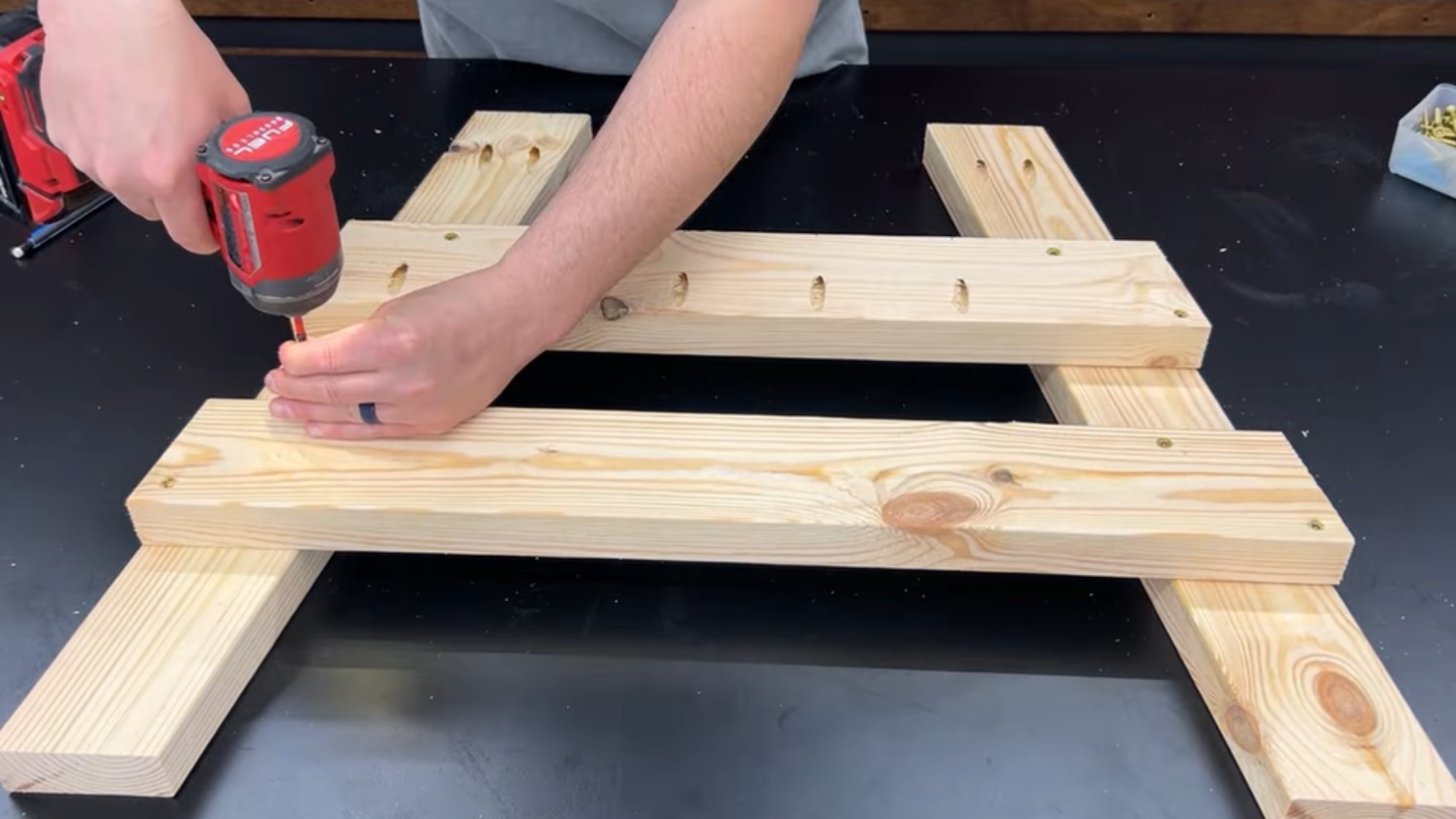 Person using a drill to join wooden planks on a black workbench for a woodworking project.