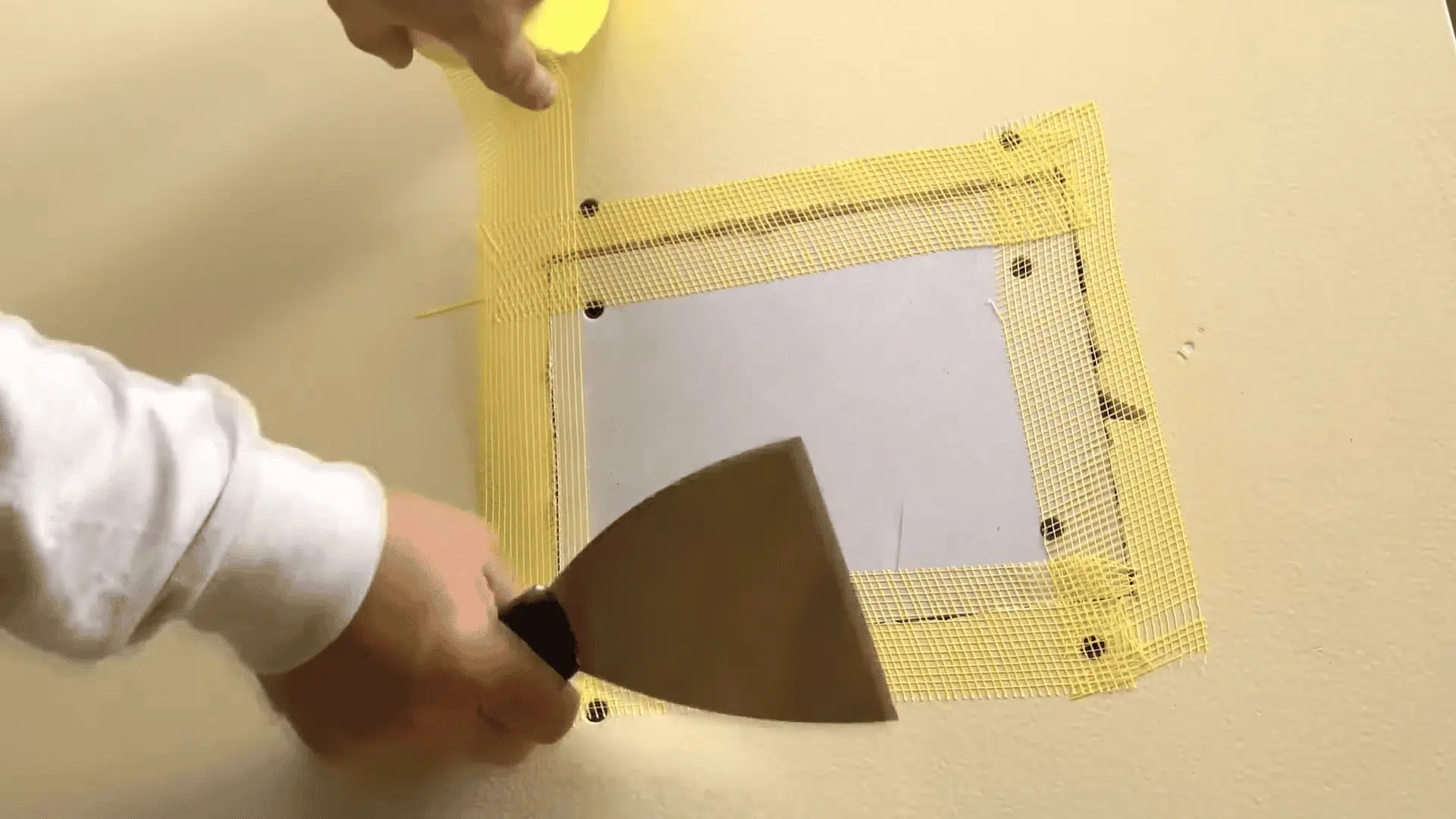 Self-adhesive yellow mesh tape is applied over the seams of a new drywall patch before mudding.