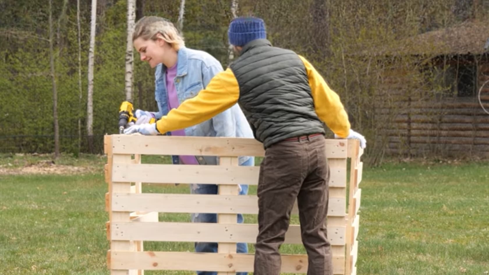 Two people assembling a wooden pallet structure outdoors on grass with tools visible