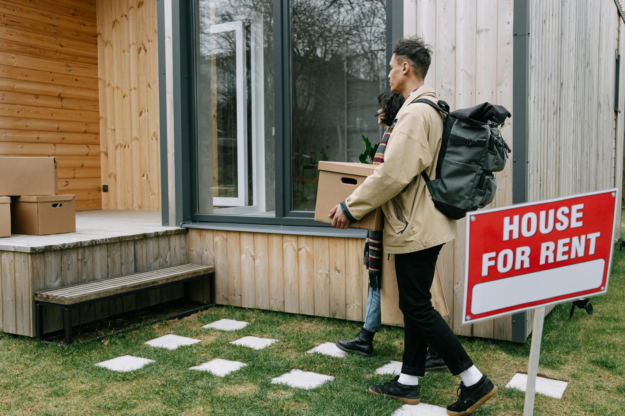 Man carrying cardboard box into wooden house with house for rent sign in yard