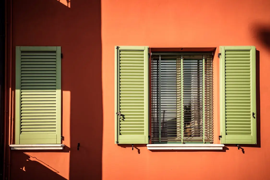 Green window shutters on bright orange wall with sunlight casting shadows