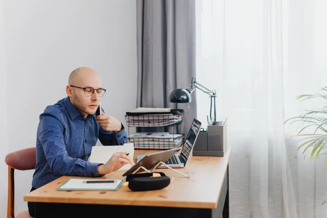 Man in blue shirt working at desk with laptop, notebook, and stack of papers in home office