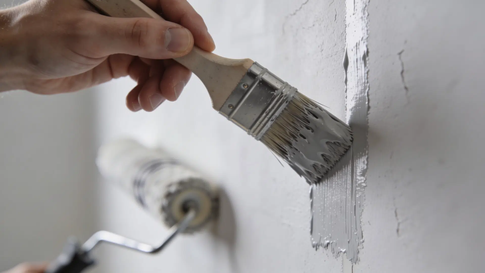 A hand applying primer to the edges of a wall with a brush, while a roller covers the flat areas.