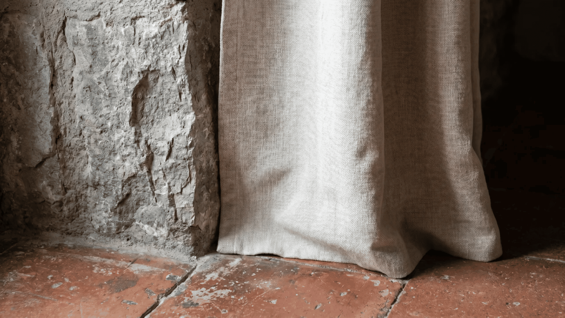 Rough stone wall beside a natural linen curtain above a worn terracotta tile floor.