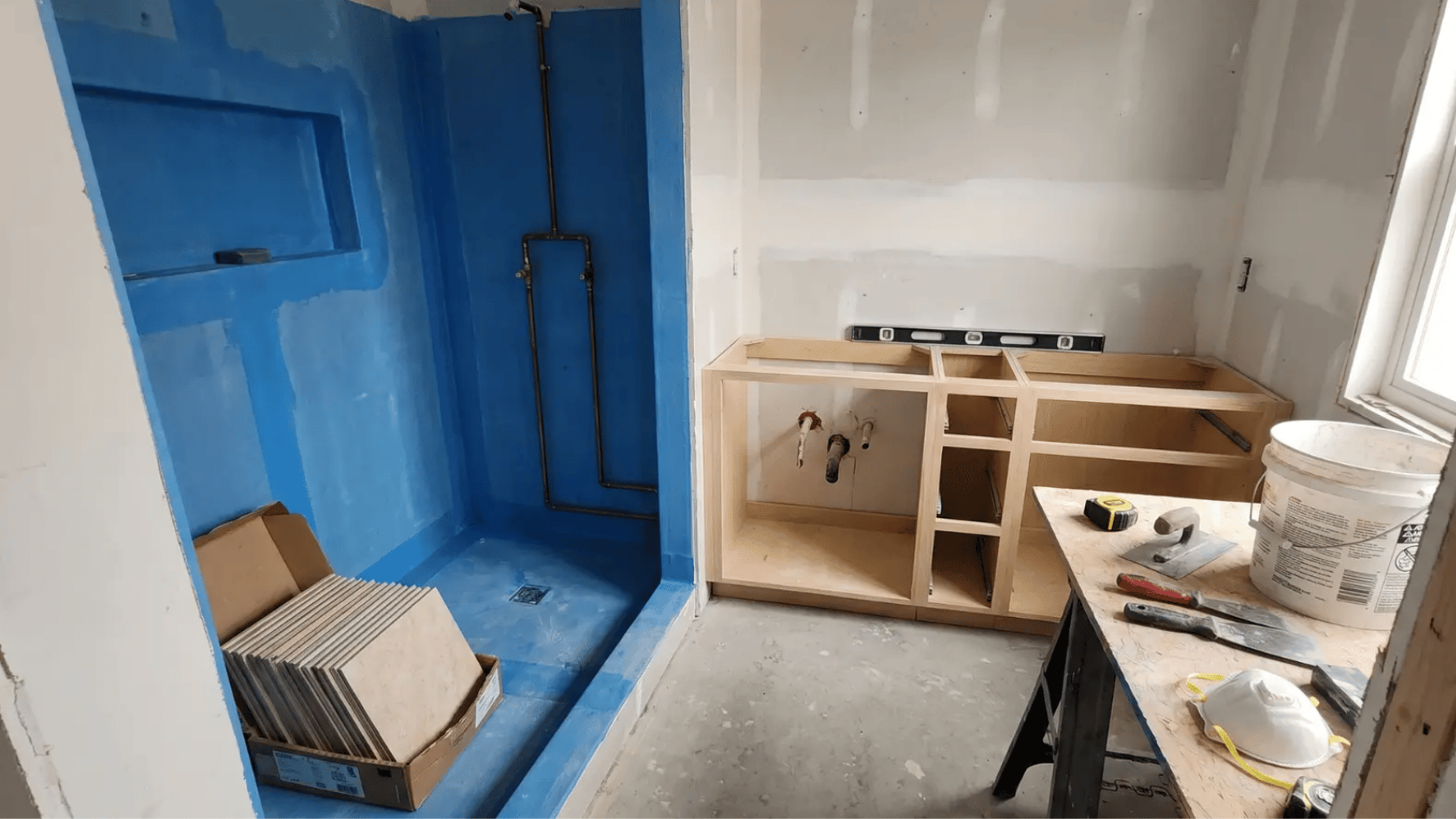 Bathroom under renovation with blue waterproofing in shower, cabinets framed, tiles stacked, and tools on a table