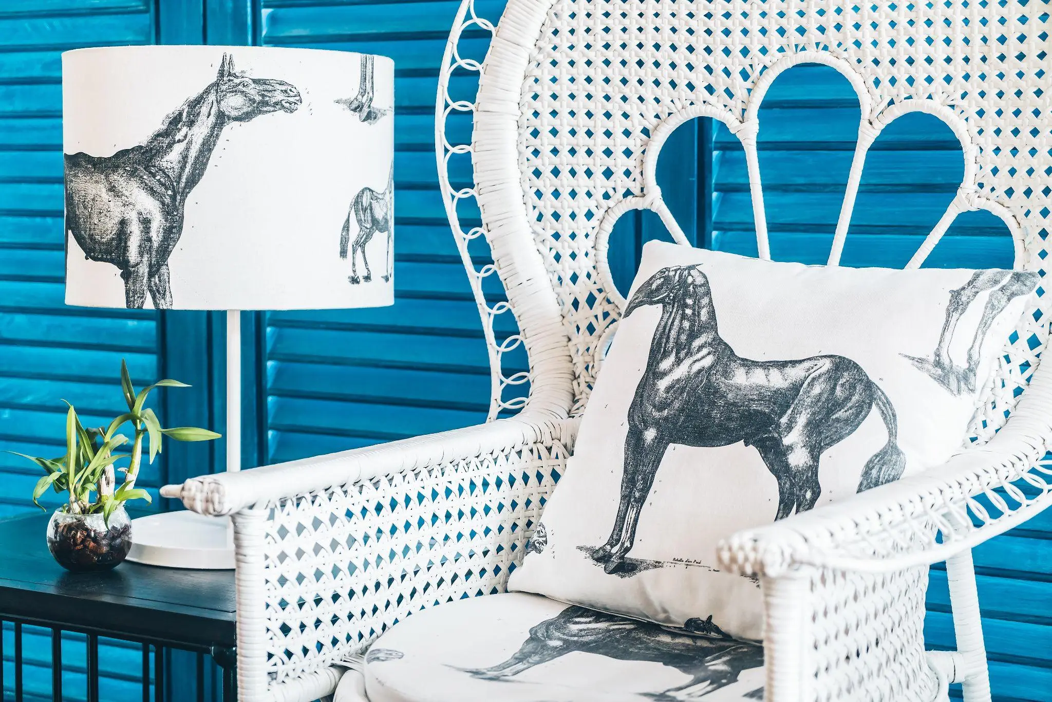 White wicker chair with horse-patterned cushions and matching lampshade in front of blue shutters