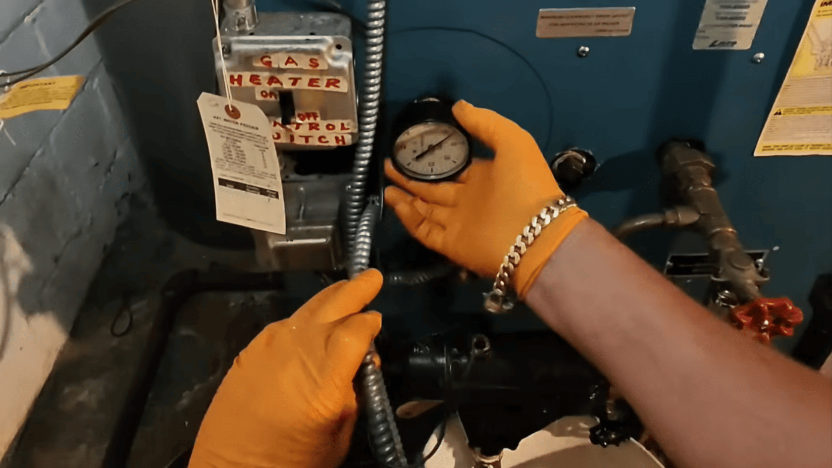 Alt Text:  Hands wearing orange gloves adjusting a boiler pressure gauge with pipes and controls visible.