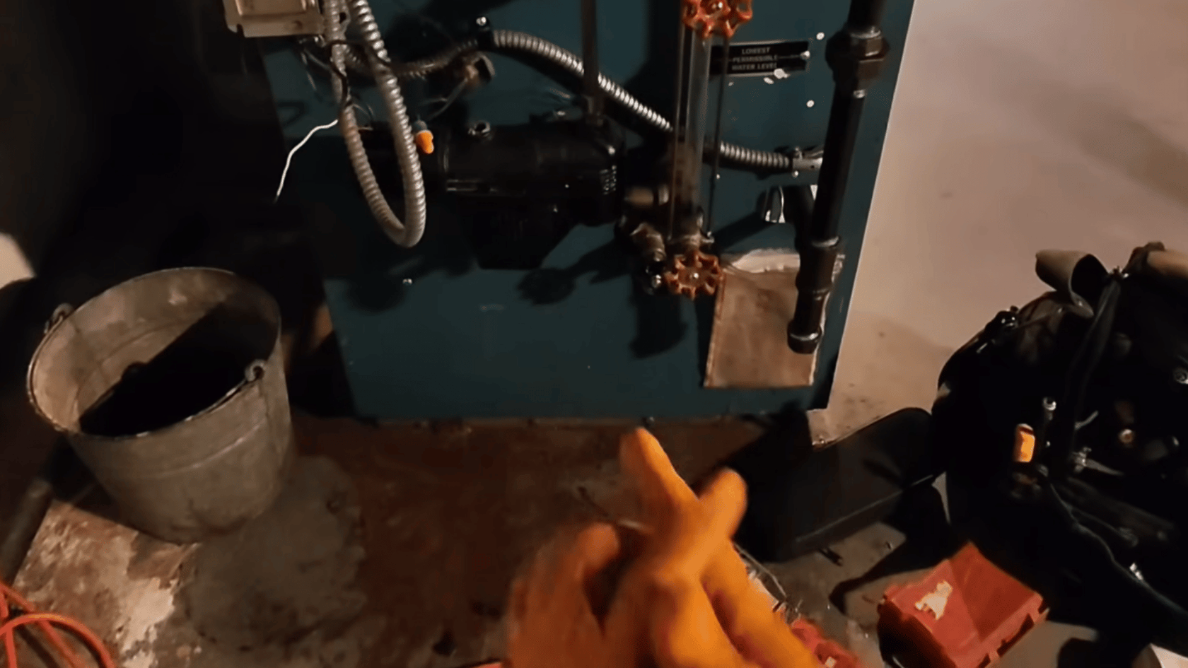Hands wearing orange gloves working on a boiler component, with pipes and boiler parts visible around.