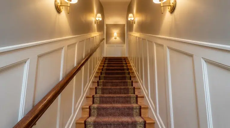 Wooden staircase with red carpet runner and white wainscoting under warm lighting sconces
