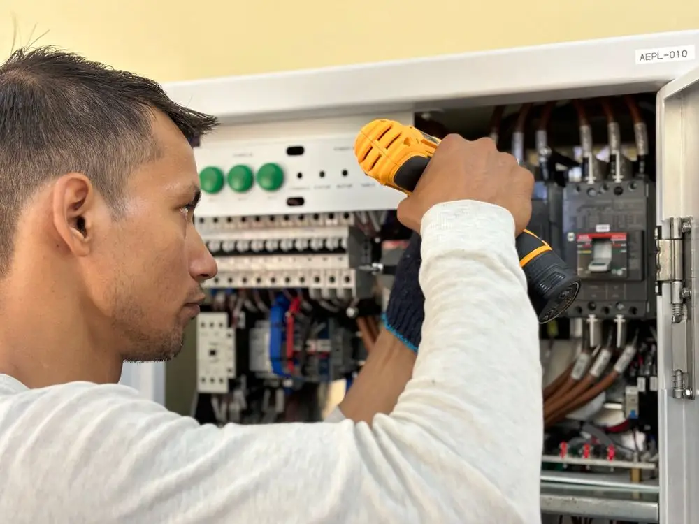 Electrician using drill to work on a circuit breaker panel in well-lit room