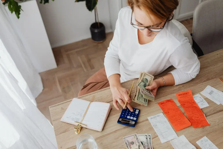 Person counting dollar bills using calculator at wooden desk with receipts and notebook
